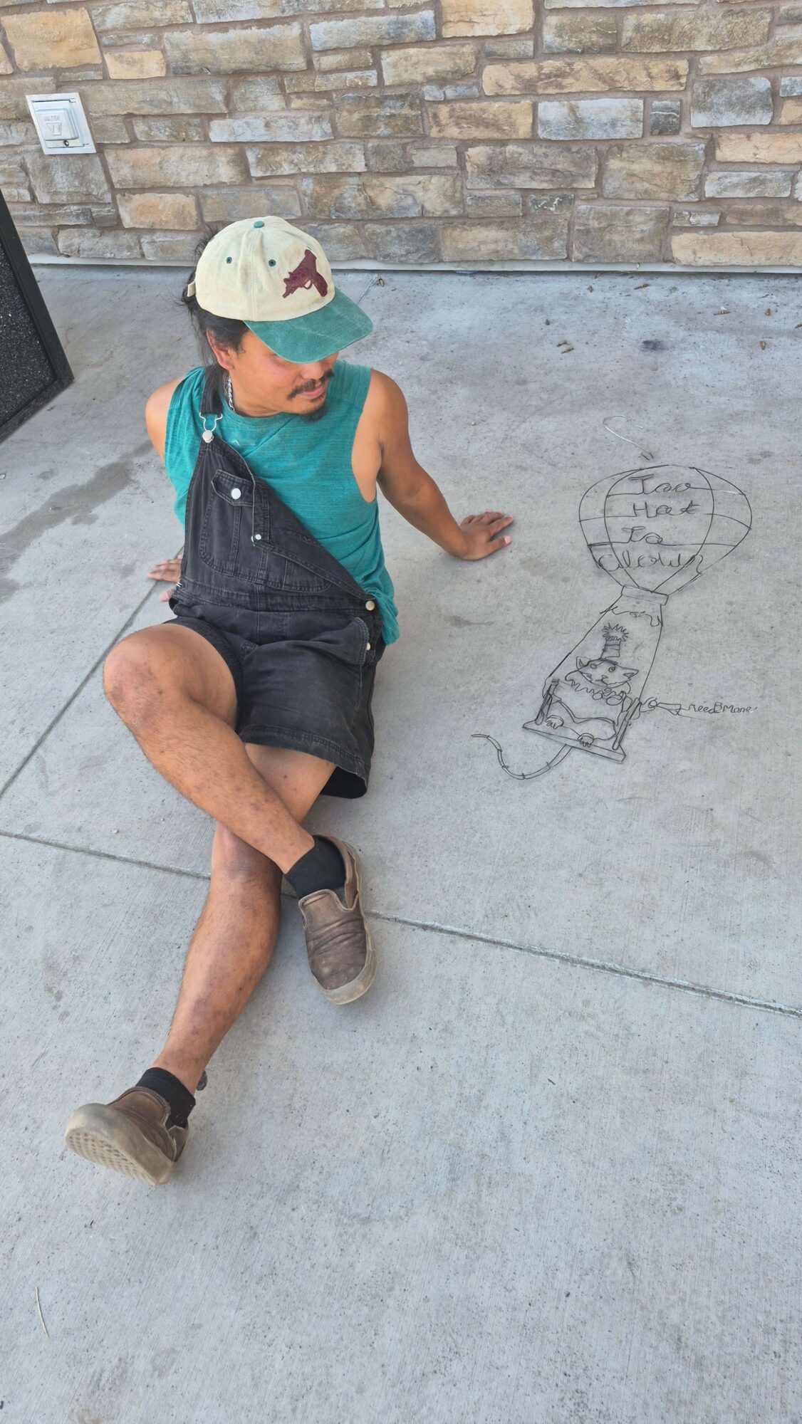 Person lying on the ground wearing a helmet, teal shirt, and black shorts, with a brick wall behind and concrete surface.