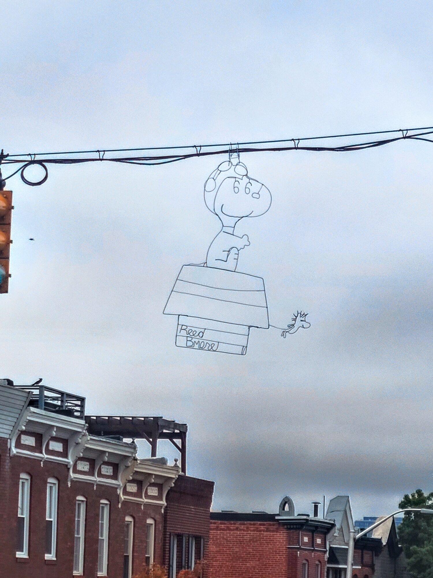 A wire sculpture of a cartoon character with a large head and small body, hanging in the sky above buildings.