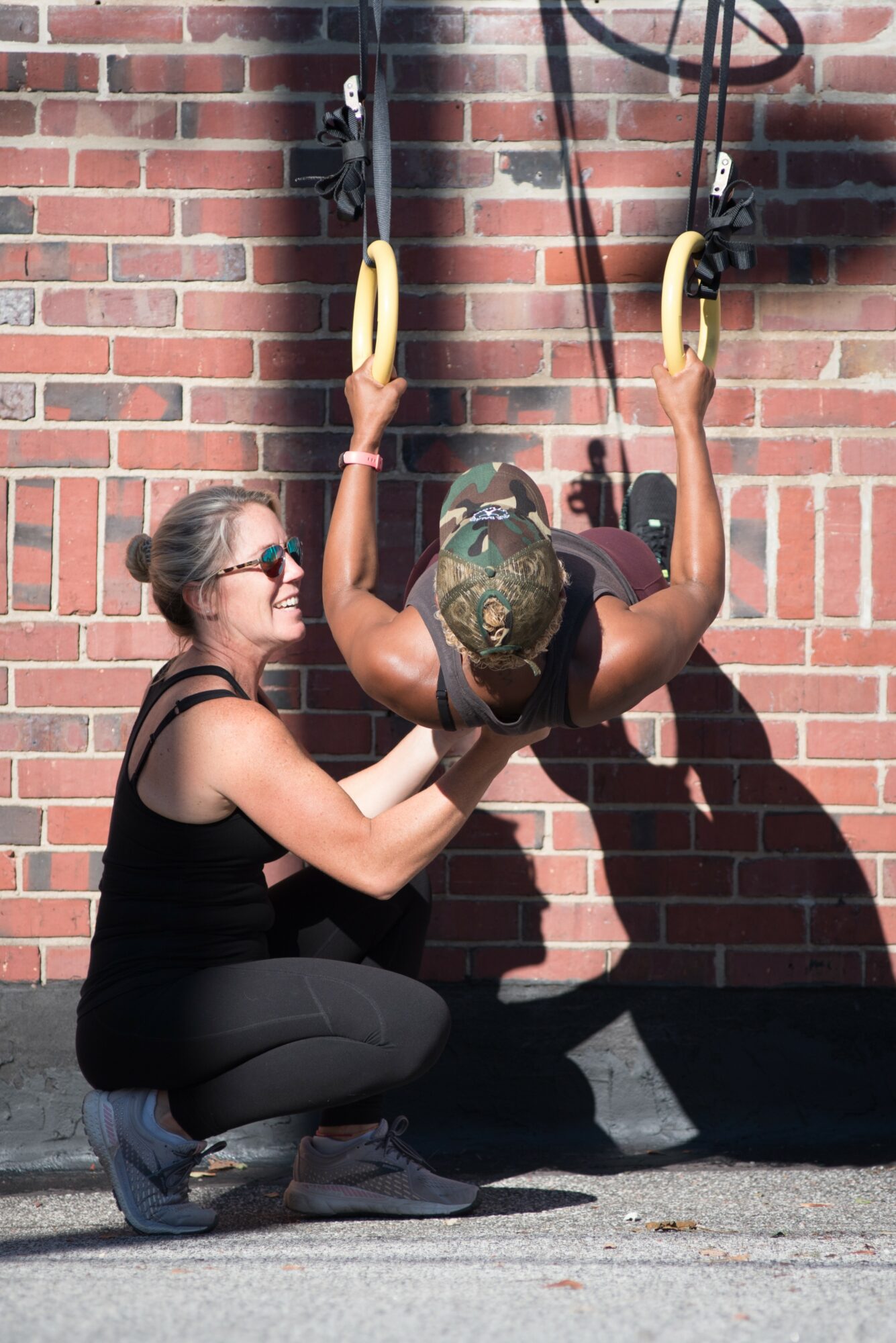 A woman assists a person hanging from gym rings against a brick wall outdoors.