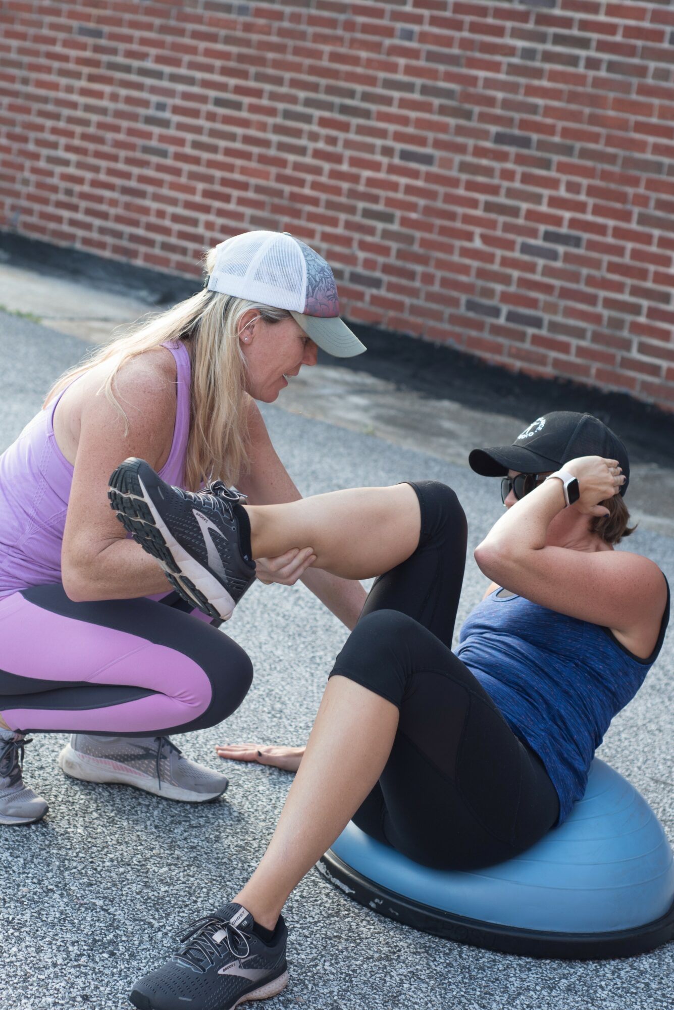 Woman in purple workout clothes assisting a woman in blue on an exercise ball outside, brick wall background.
