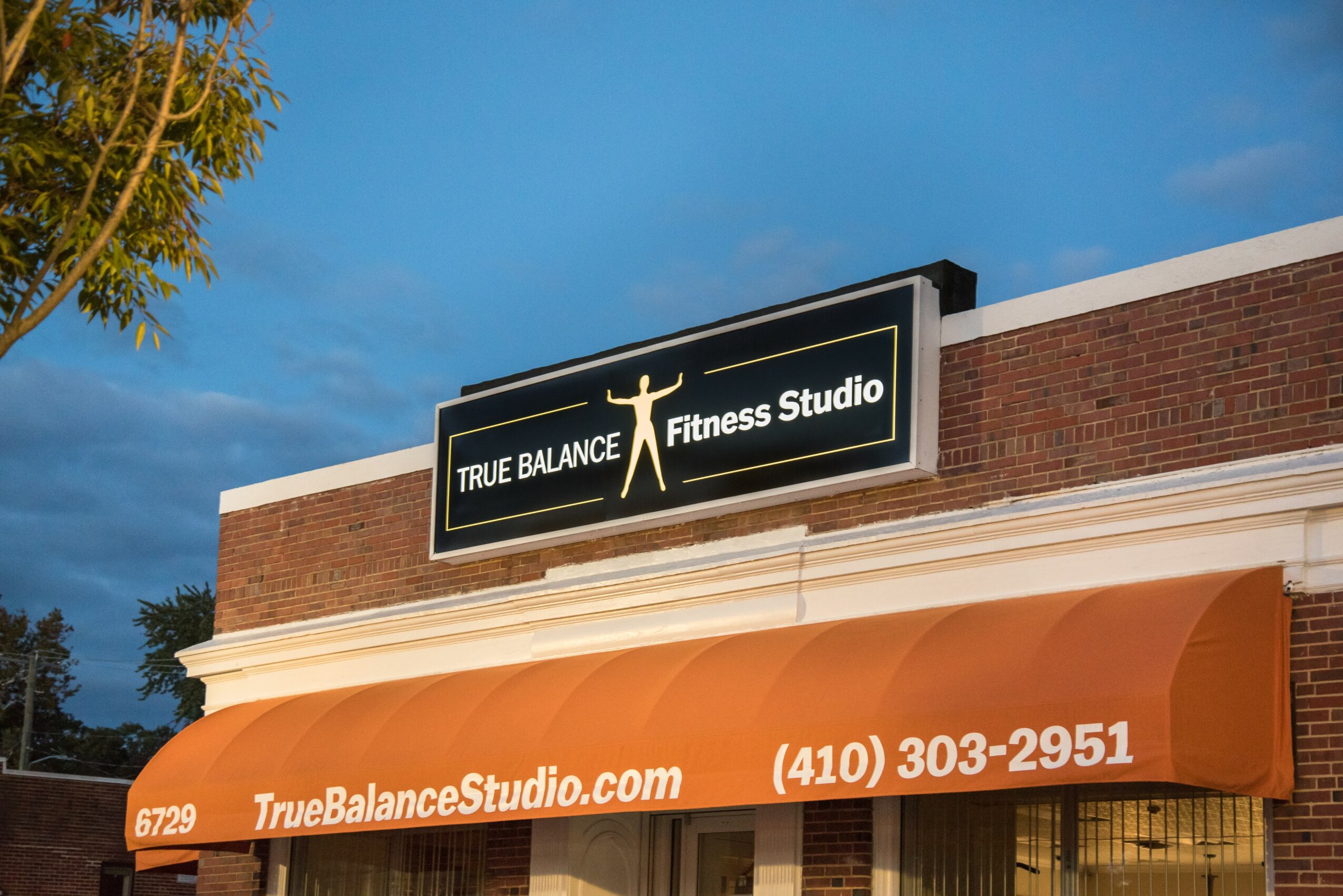 Sign for True Balance Fitness Studio on building with orange awning, blue sky background.