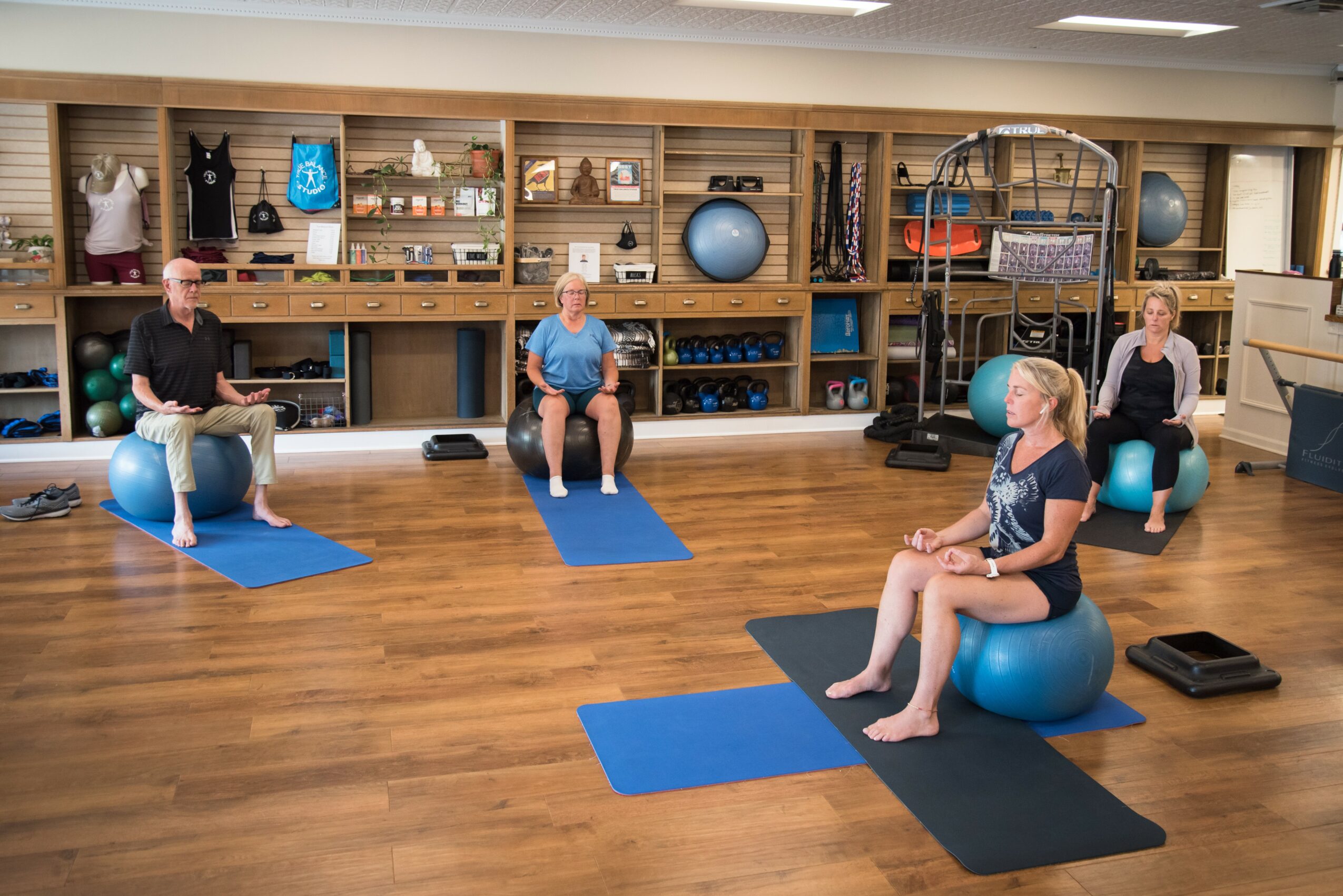 Four women seated on exercise balls and mats in a fitness studio with shelves of equipment behind them.