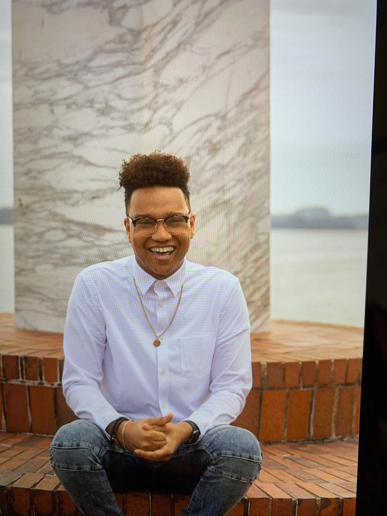 Man with glasses, smiling, wearing a white shirt, sitting on a brick ledge outside, with a marble wall behind him.
