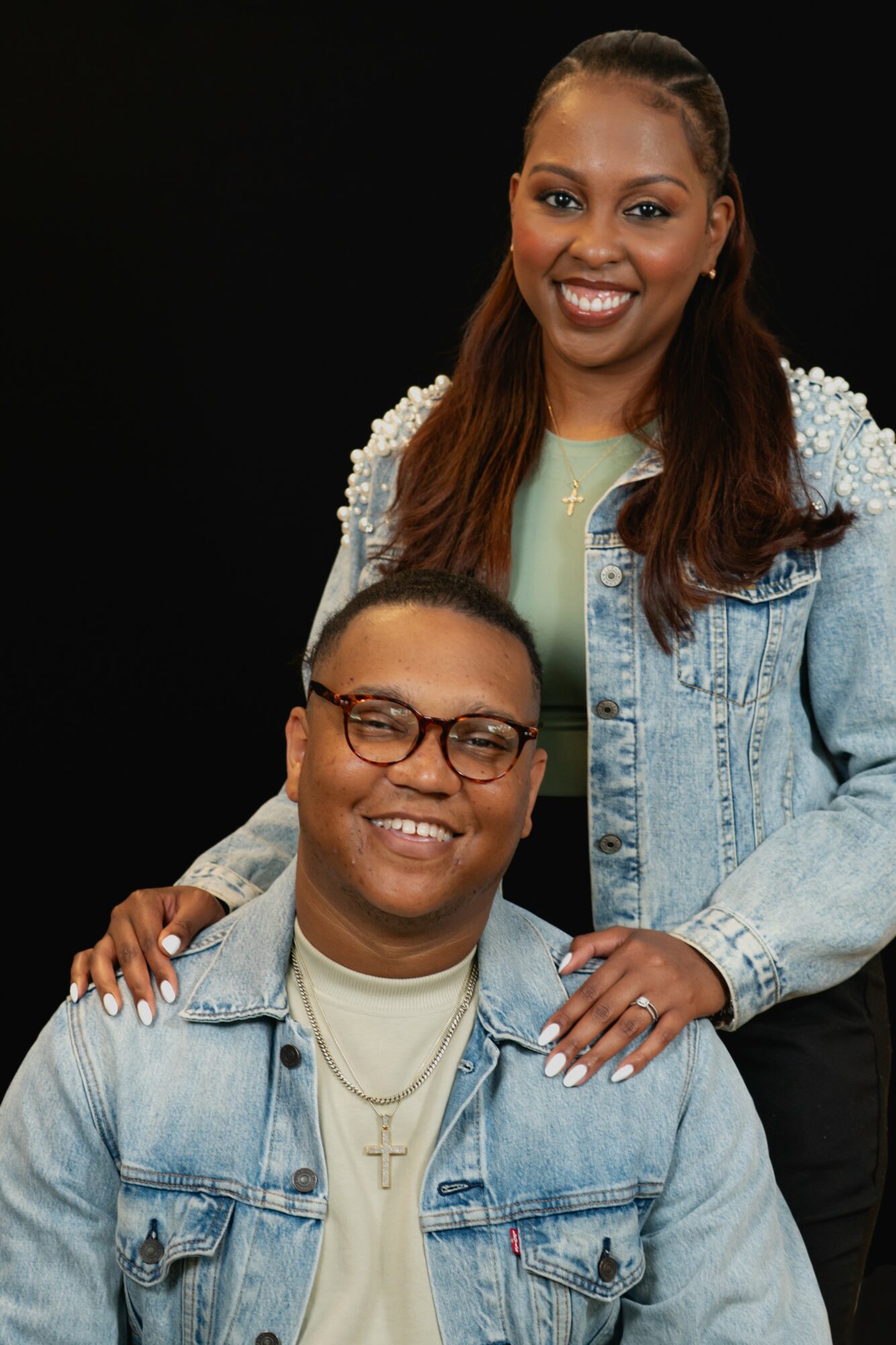 A smiling woman with long hair and a denim jacket stands behind a smiling man with glasses and a denim jacket, against a black background.