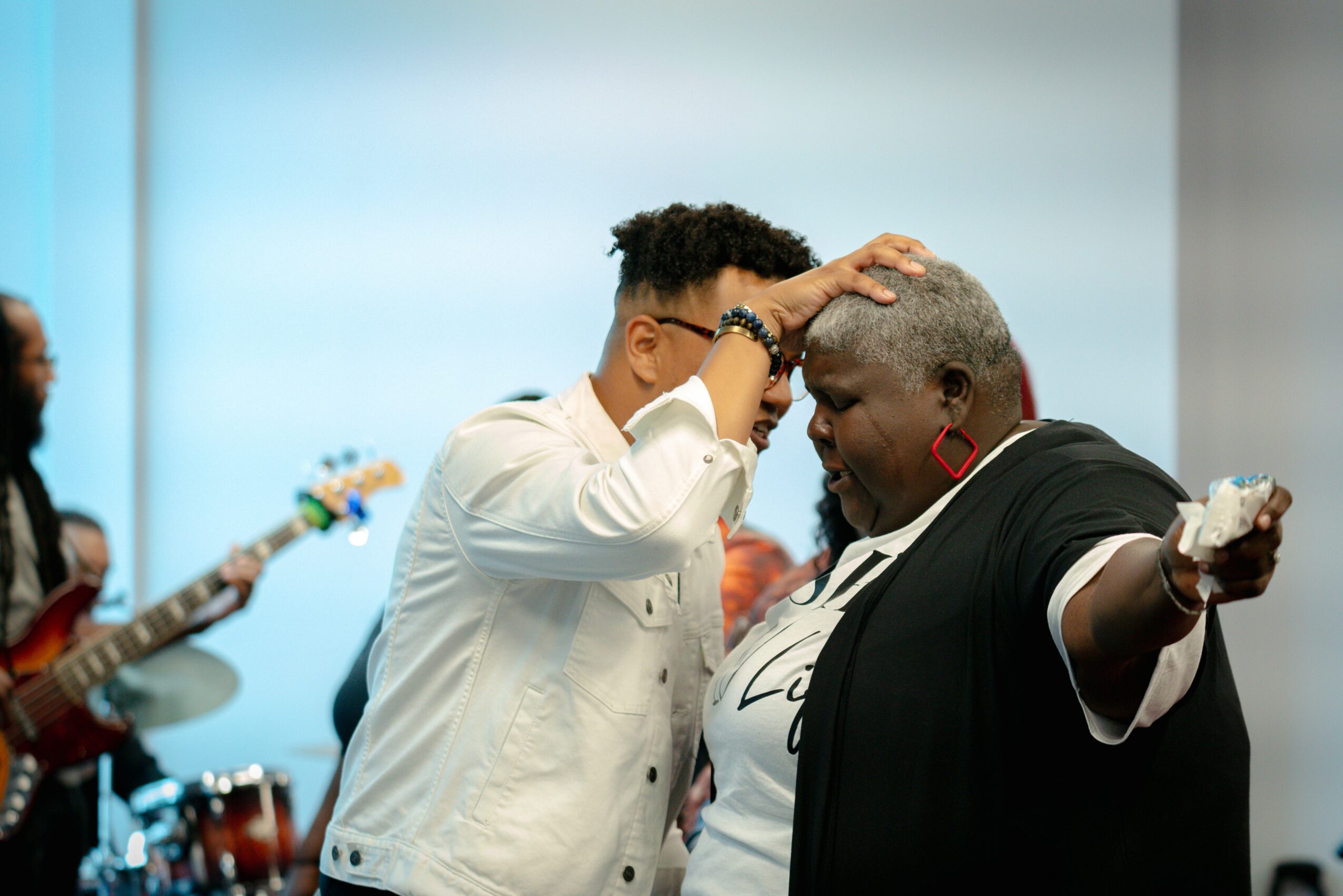 Two women share a moment, one touching the other's head, with musicians playing in the background.