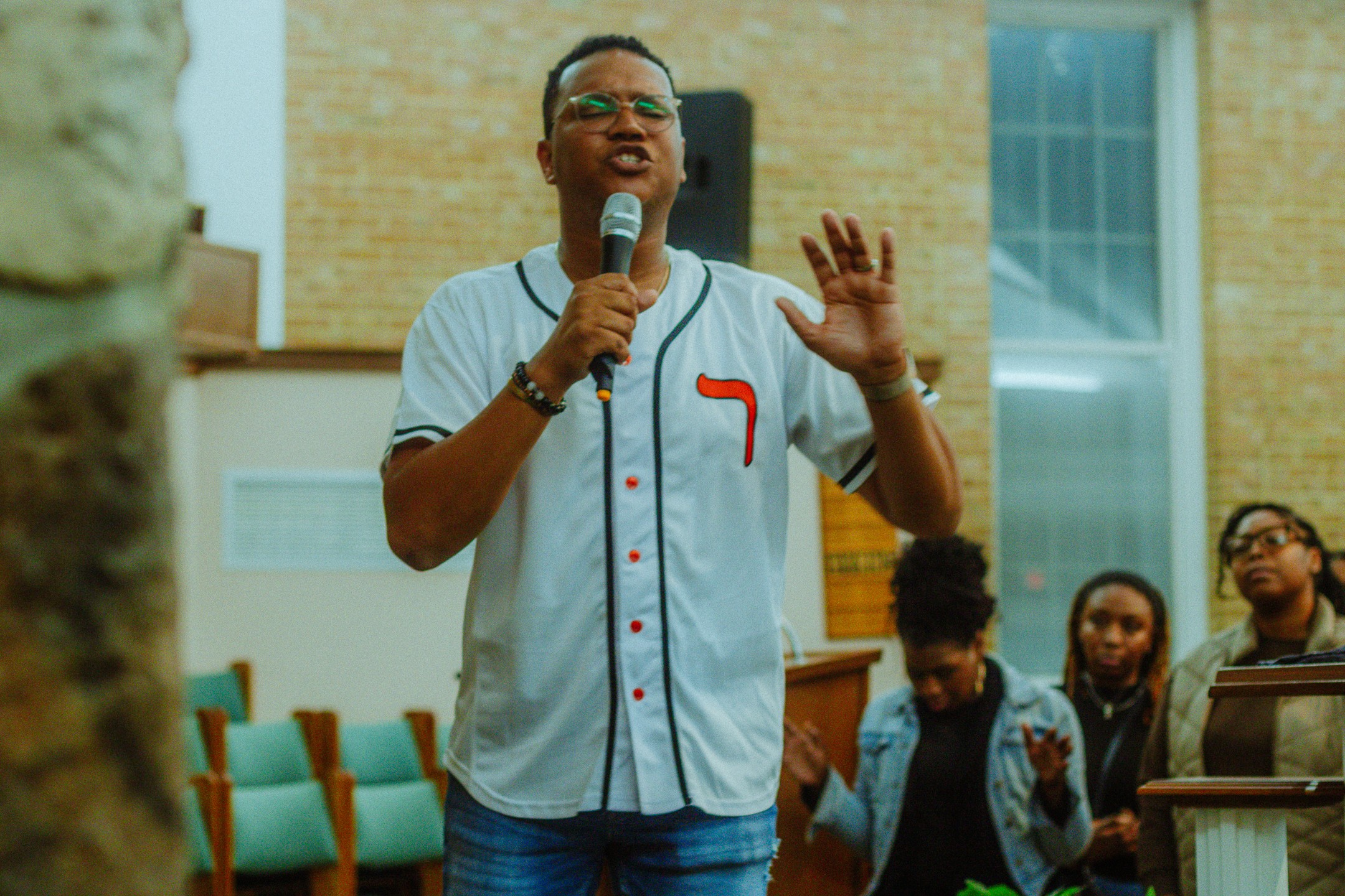 Person speaking into microphone, gesturing with hand, audience listening attentively, indoor setting with brick wall and large window