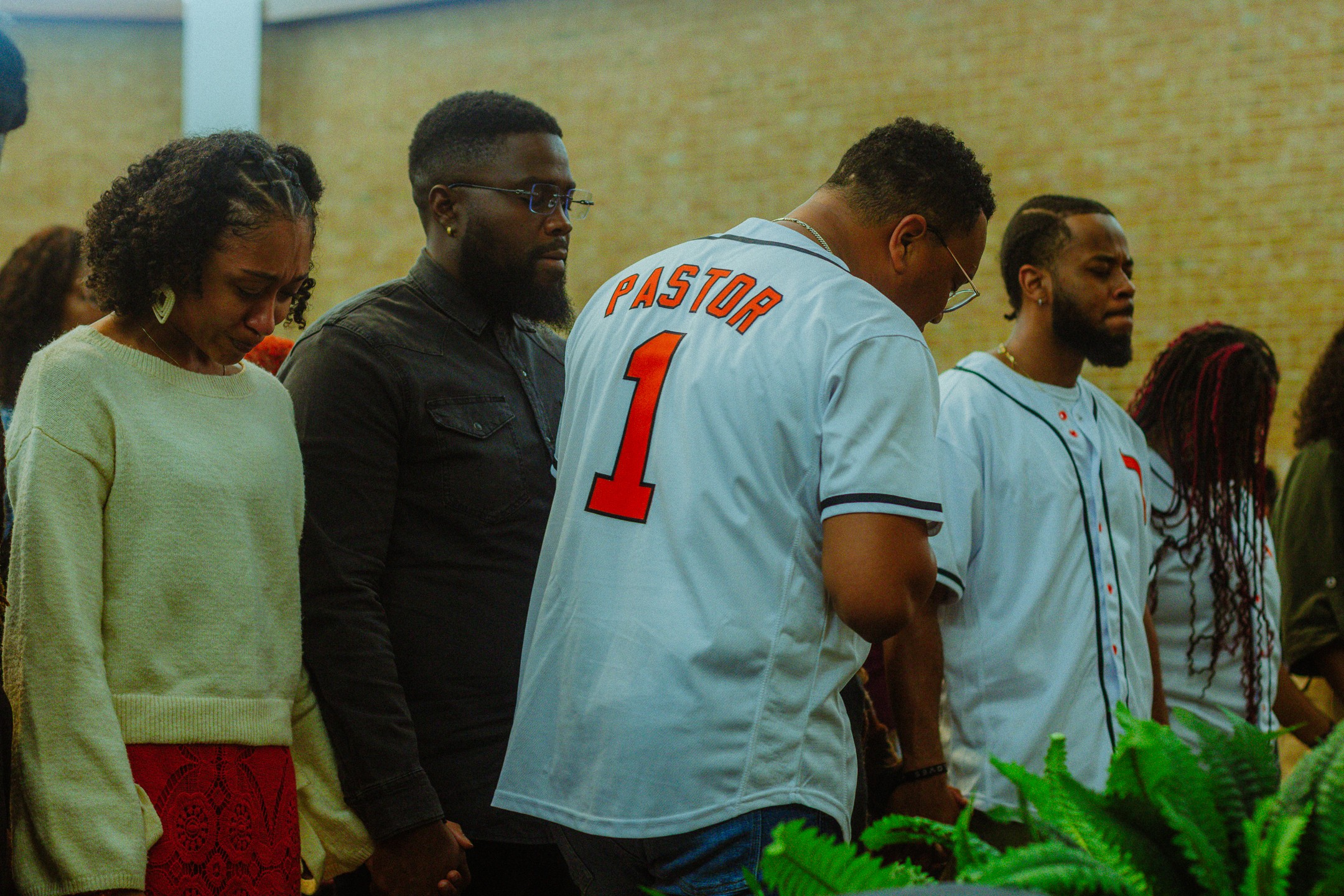 Group of people standing indoors, some looking down, with a brick wall background and green plants in the foreground.