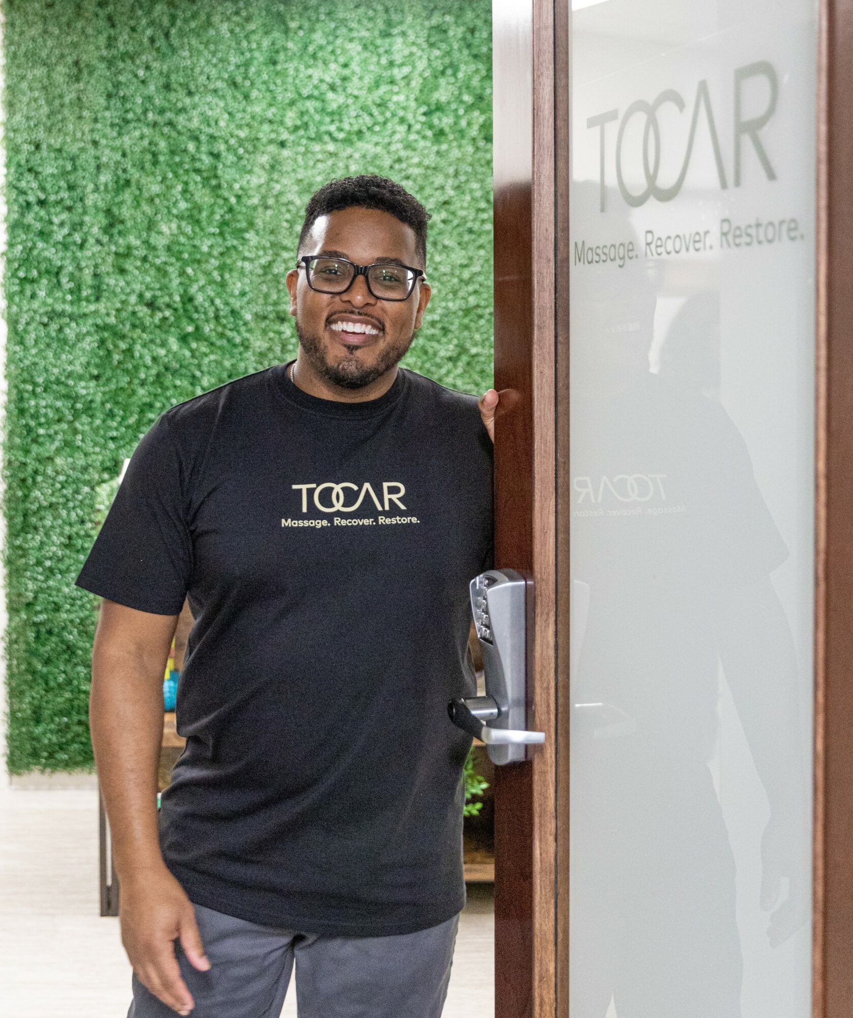 Smiling man in black T-shirt standing next to a door with frosted glass, green wall background.
