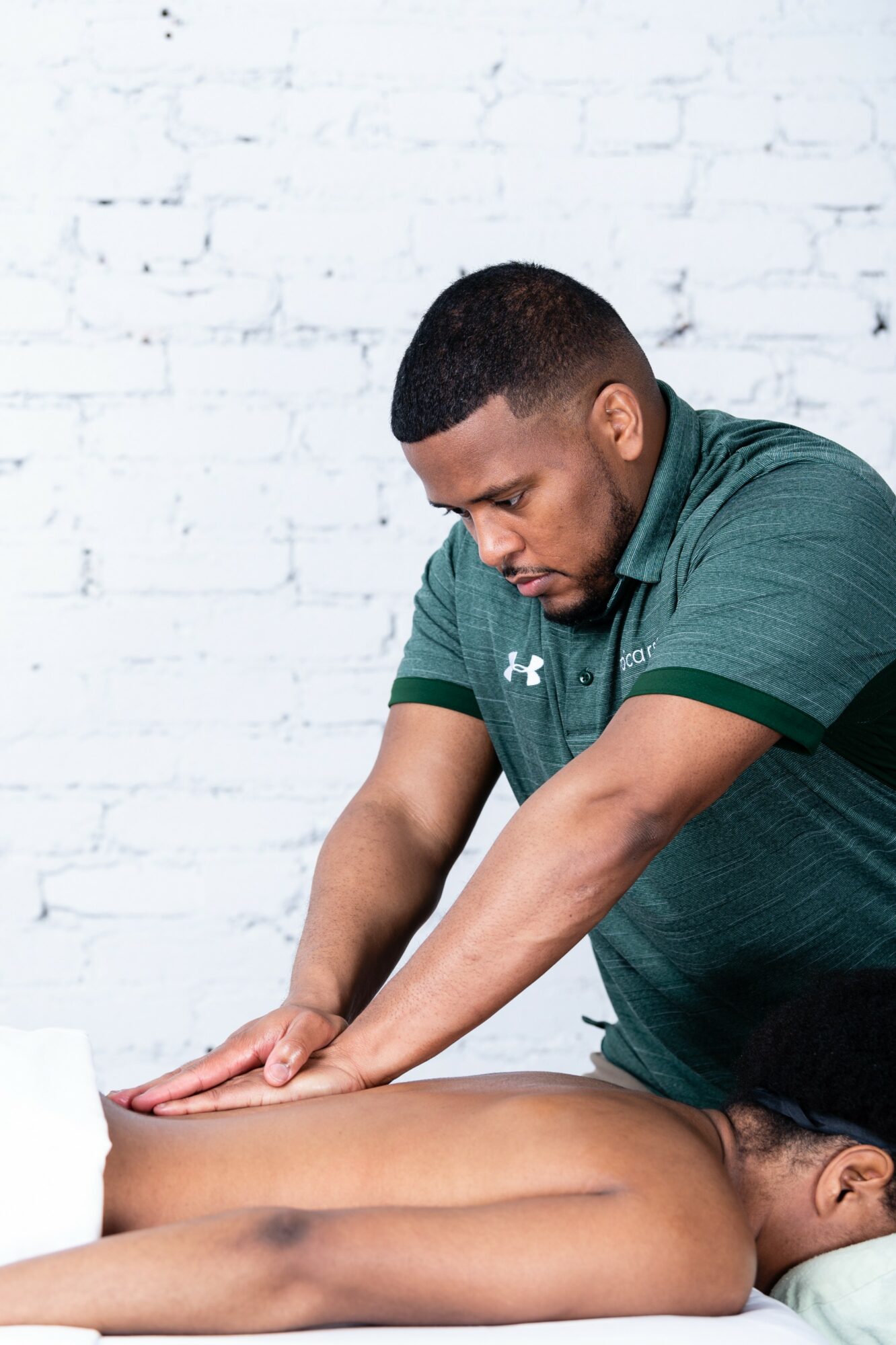 Man giving massage to a person lying face down on a massage table, white brick wall background.