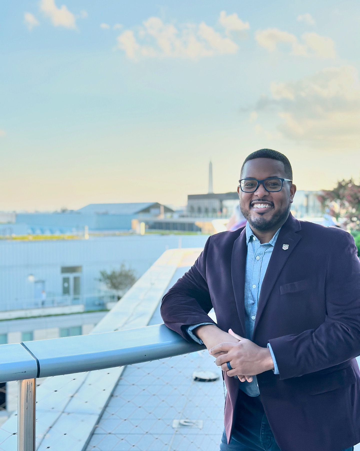 Man in a suit smiling outdoors with buildings and sky in background.