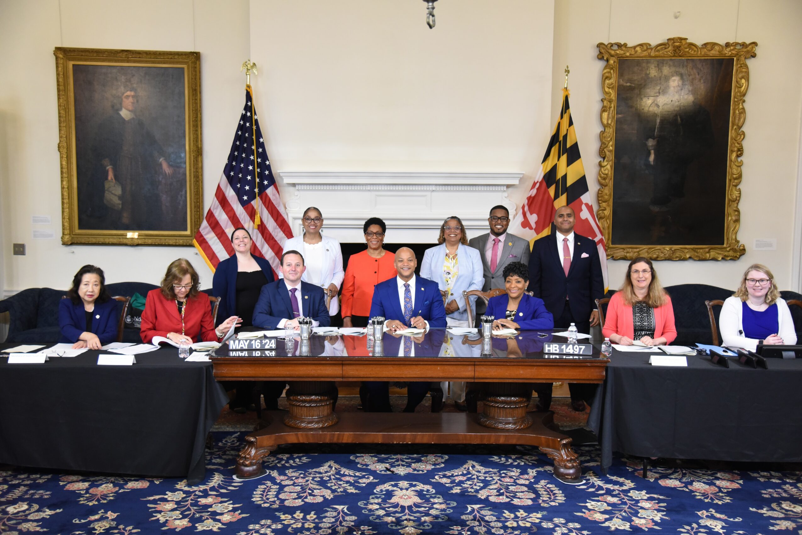 Group of people seated at a long table in a room with portraits and flags, some standing behind the table.