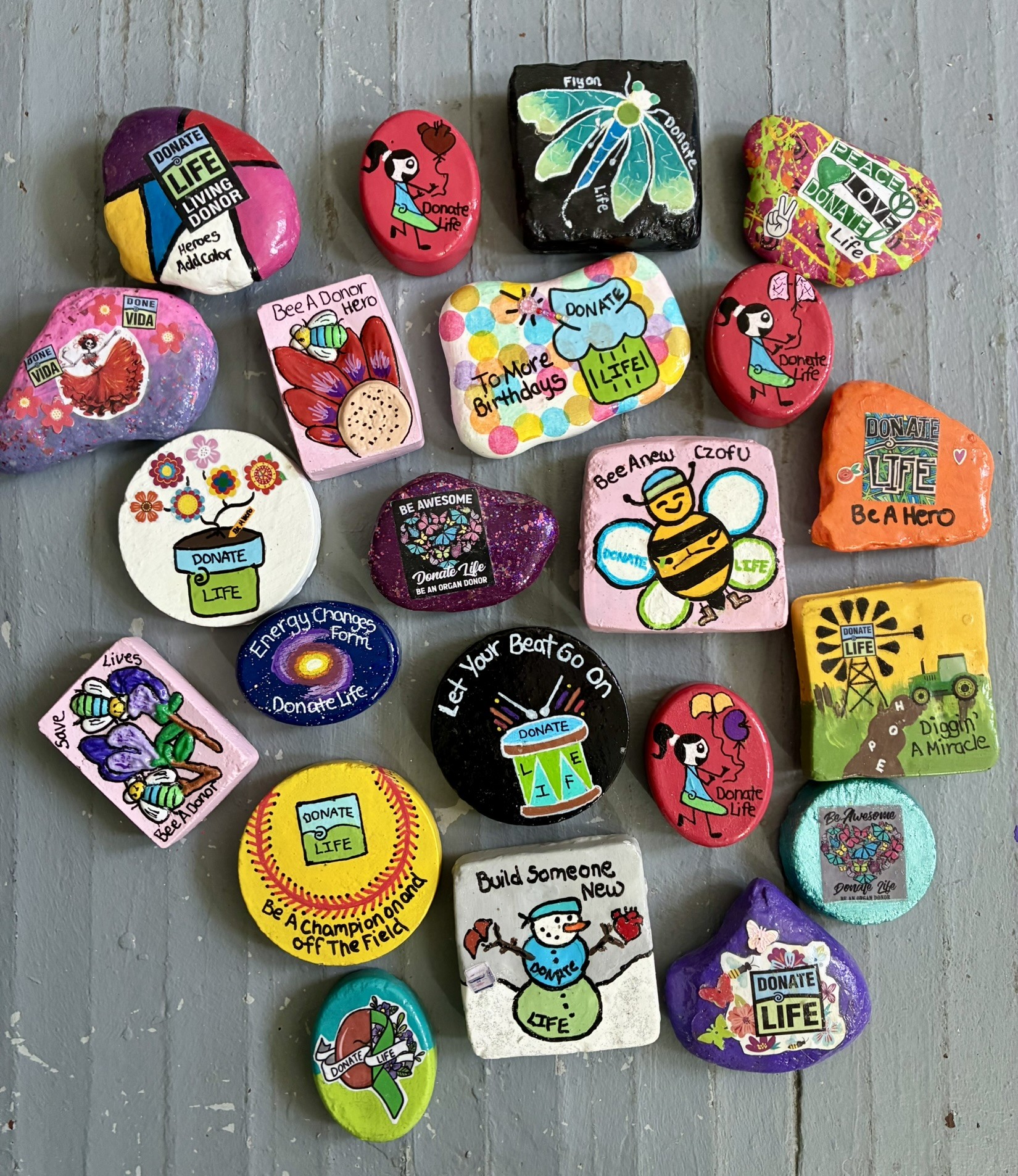 Collection of colorful decorated rocks with various designs and messages on a gray wooden surface.