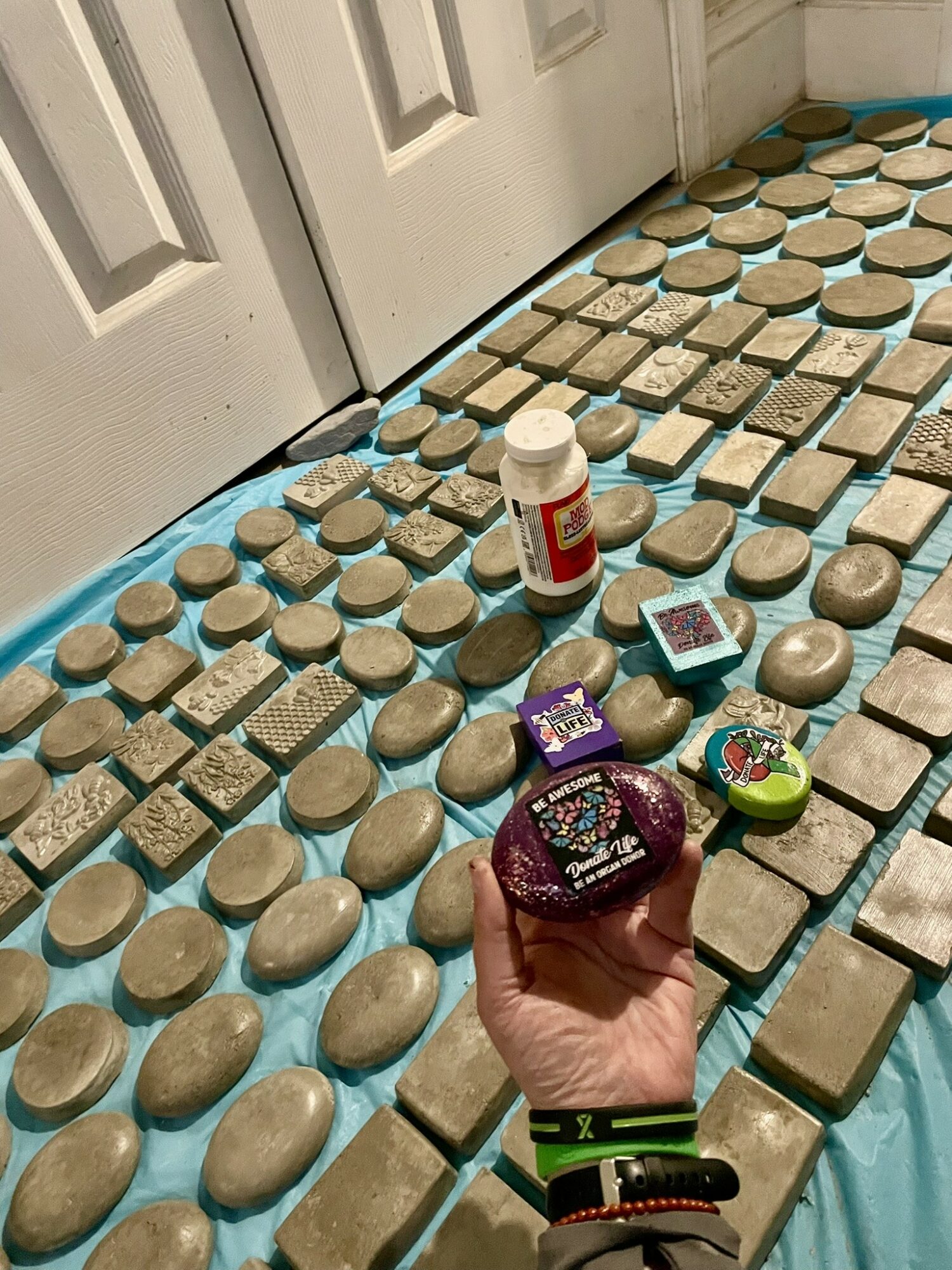 Hand holding a decorated cookie with purple icing, surrounded by various cookies, pills, and a small electronic device on the floor.
