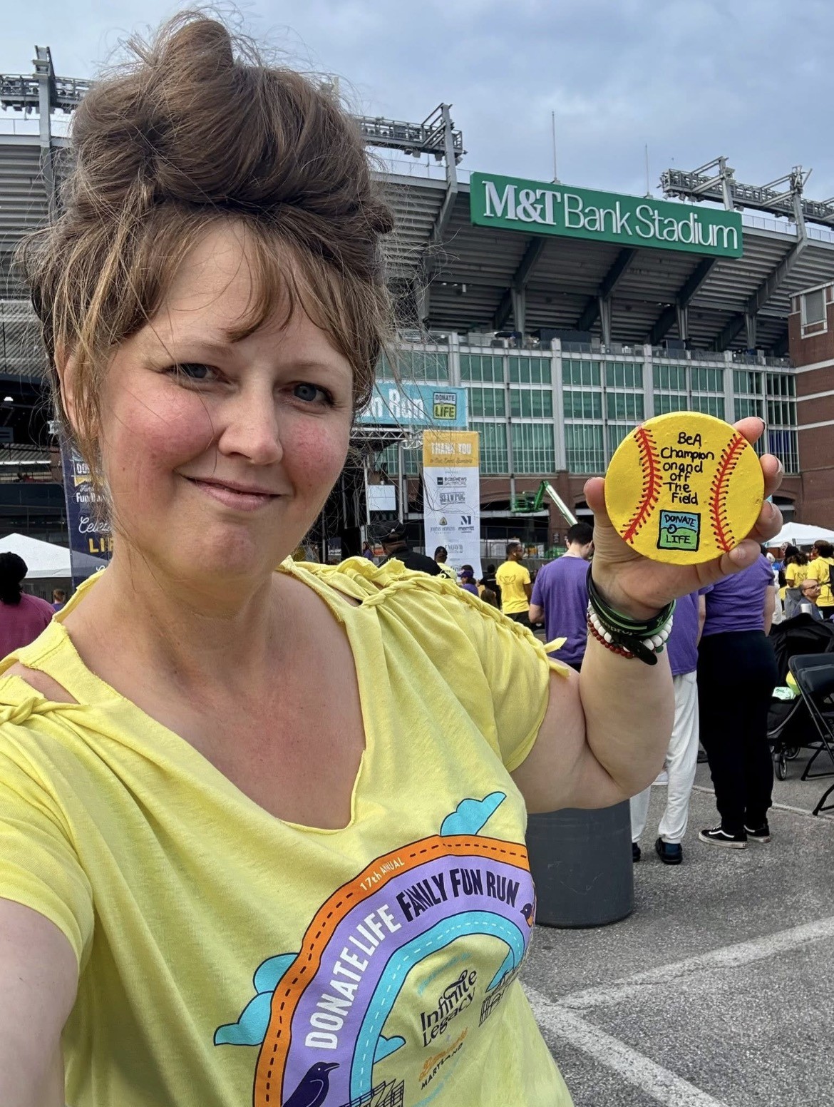 Woman holding a yellow football outside M&T Bank Stadium, smiling, with people in the background.