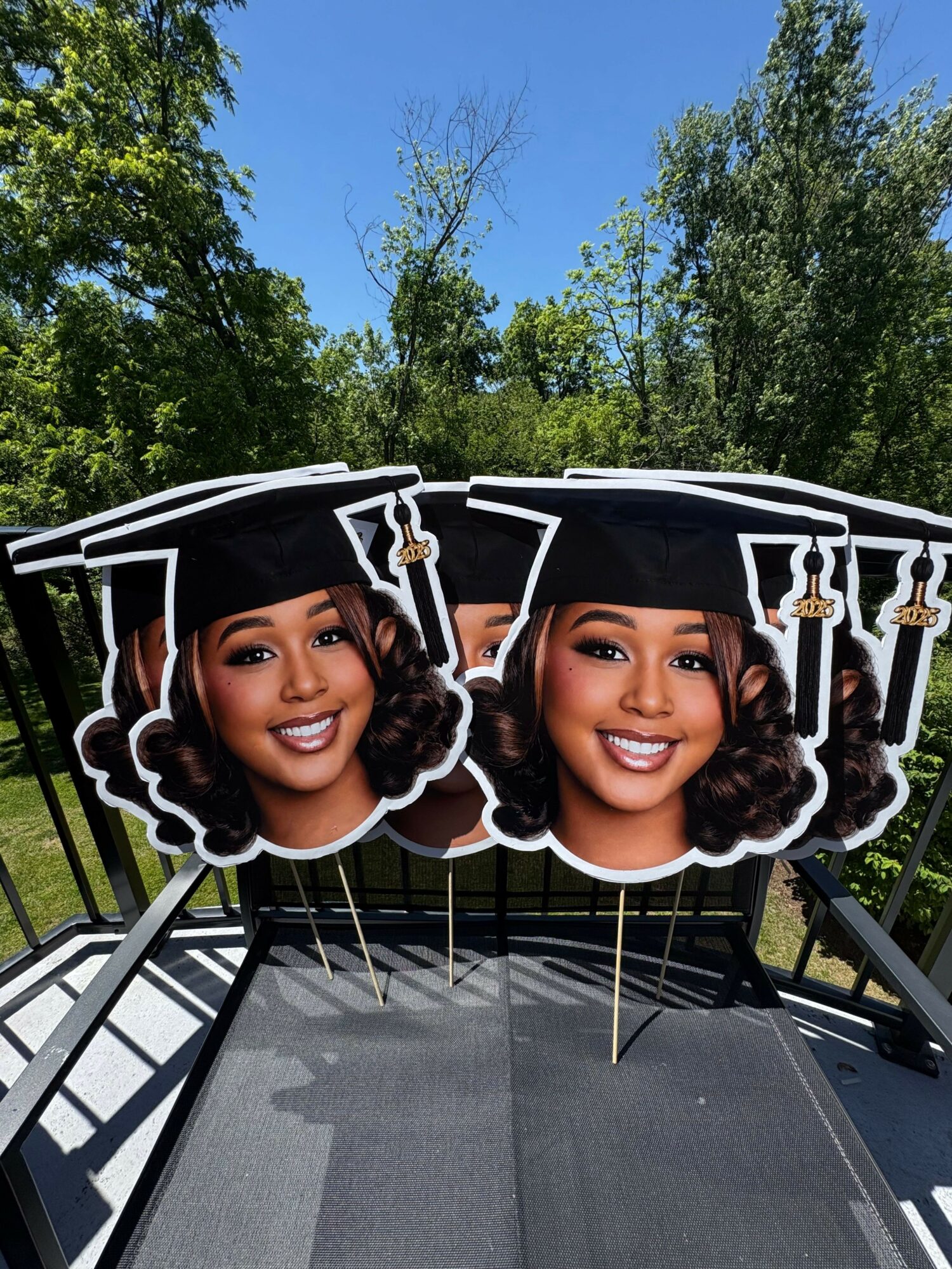 Two women with curly hair smiling, wearing black graduation caps, standing outdoors with trees and blue sky in background.