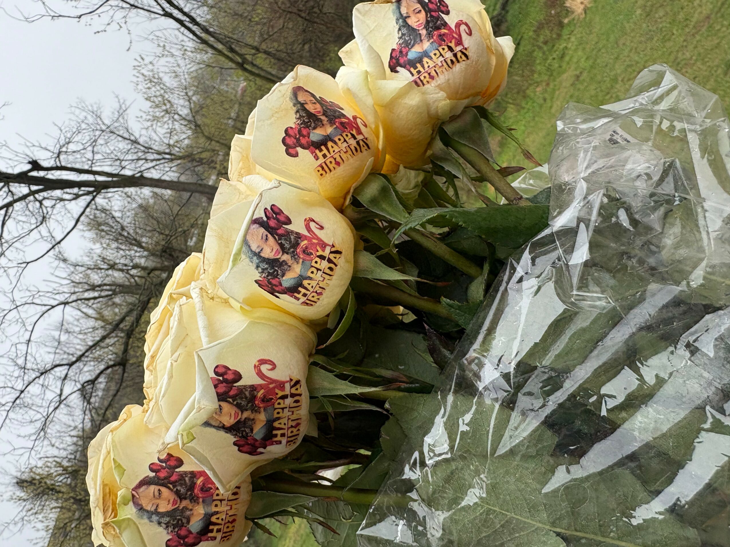 Bouquet of yellow roses with printed edible images on petals, wrapped in clear plastic, outdoors with trees and grass.