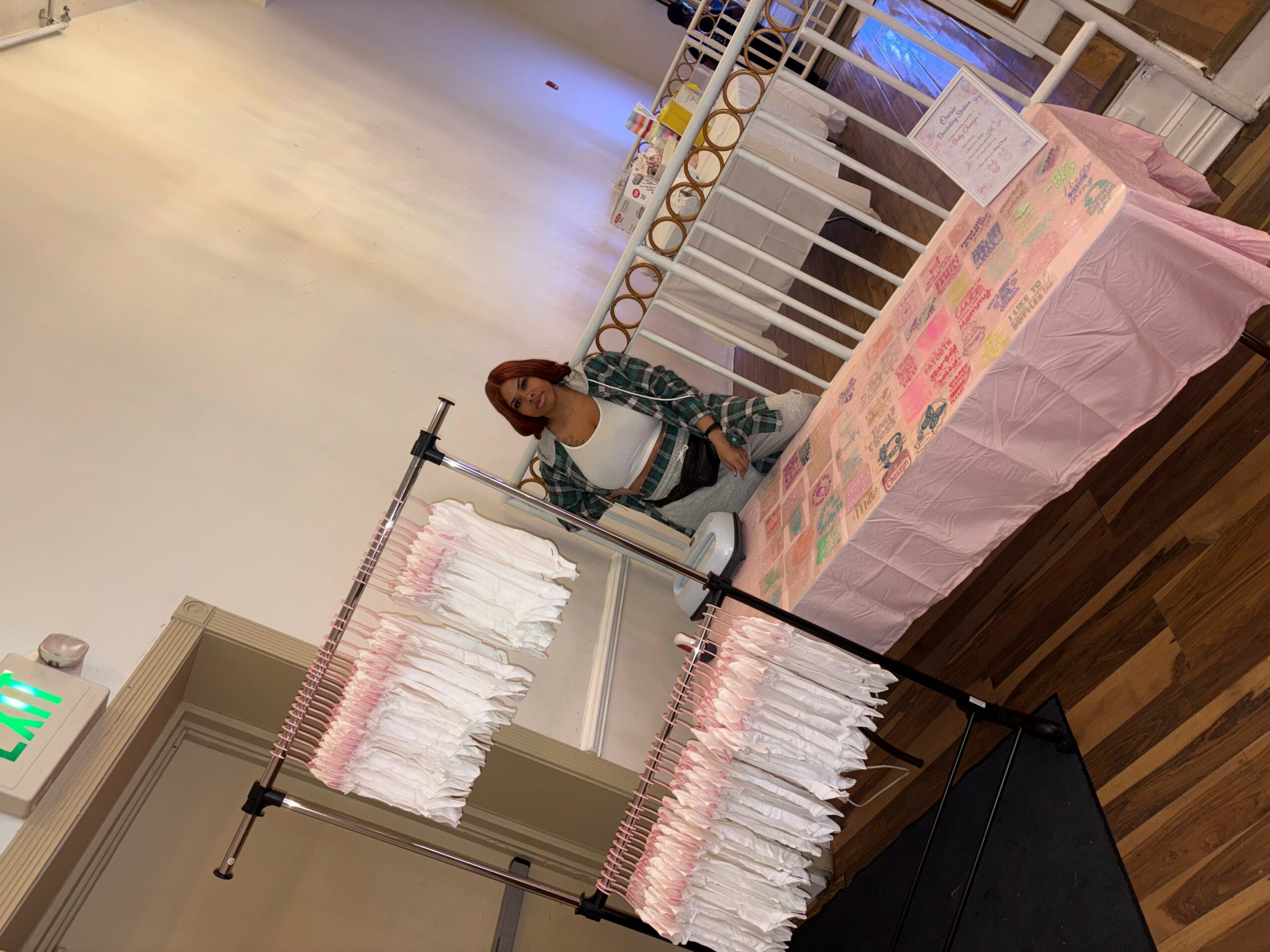 A woman stands behind a table with pink and white items, in a room with white walls and wooden flooring.
