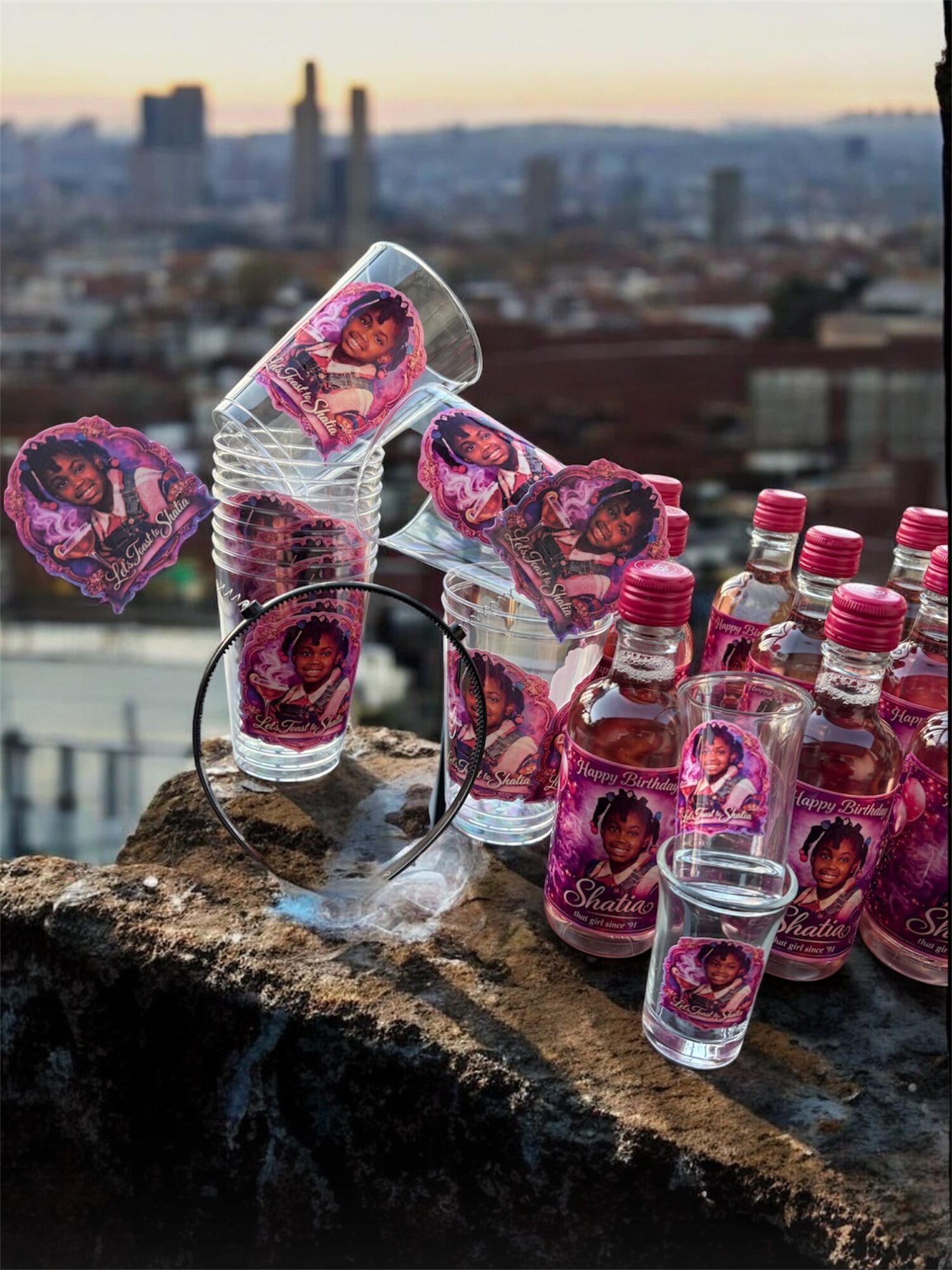 Glass bottles and cups with pink labels and a city skyline in the background on a rooftop.