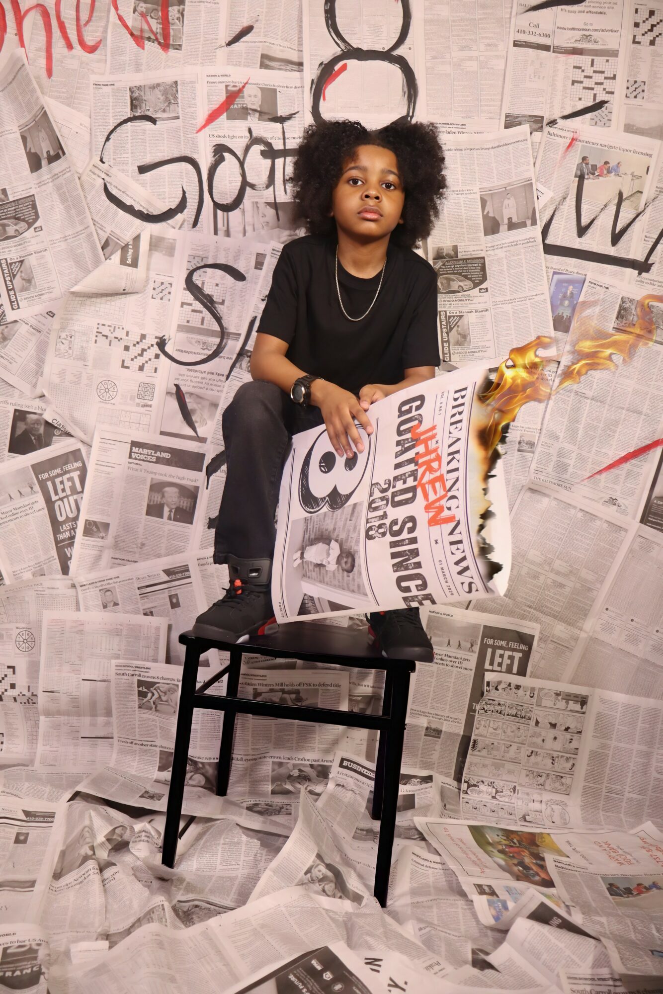 Child with curly hair sitting on a chair, surrounded by newspaper-covered walls and floor, holding a newspaper.
