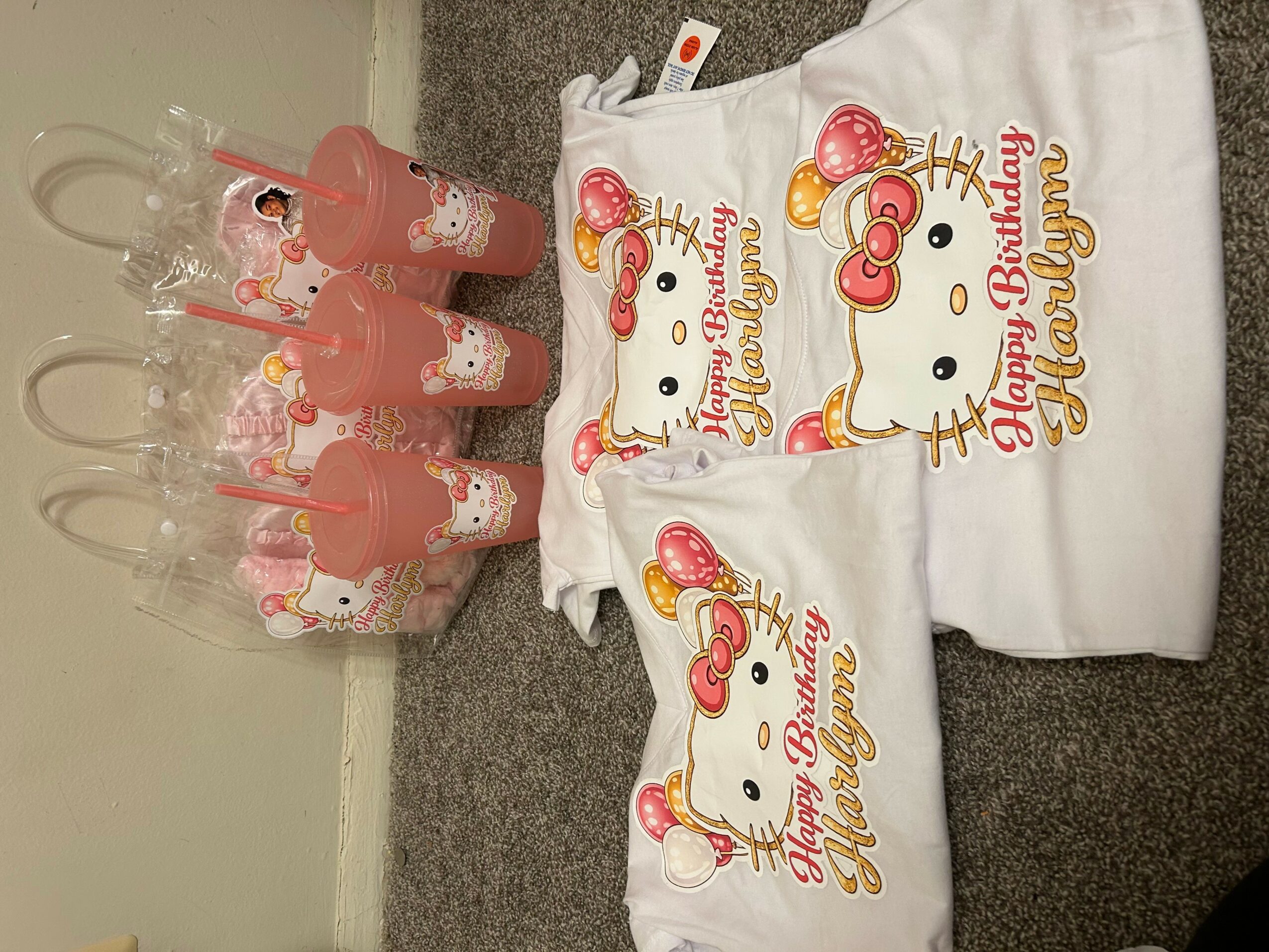Six pink bottles with Hello Kitty designs and three white T-shirts with Hello Kitty birthday graphics on a gray carpet.