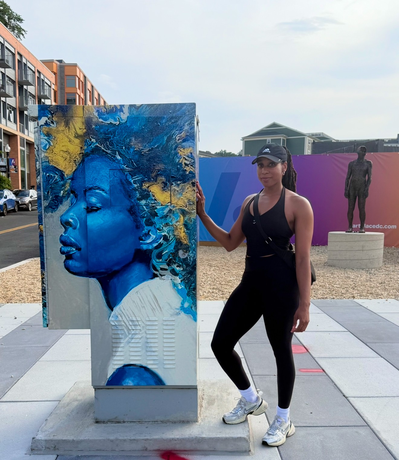 Woman in black athletic clothing and white sneakers standing next to a colorful portrait sculpture outdoors.