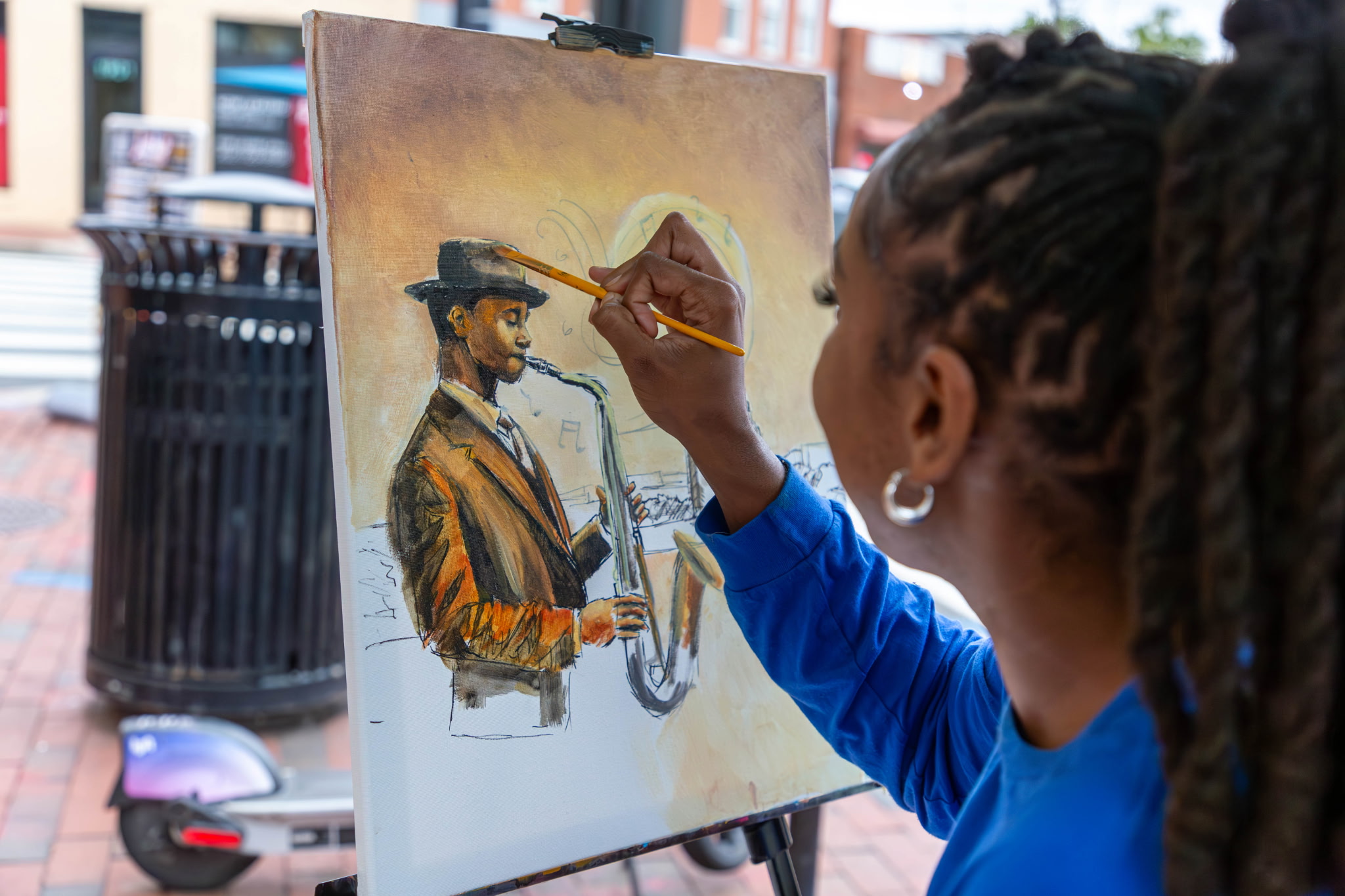 Person with braided hair painting a woman in a hat on a canvas outdoors.