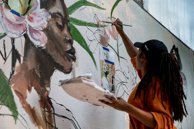 Person painting a large mural of a woman's face with flowers, using a brush and palette, indoors.
