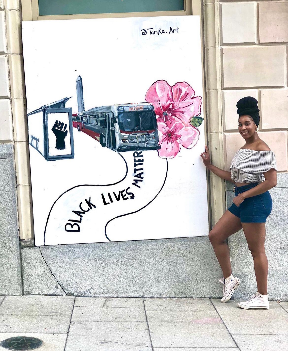 Young woman with a bun hairstyle, wearing a white off-shoulder top, denim shorts, and white sneakers, standing next to a mural.