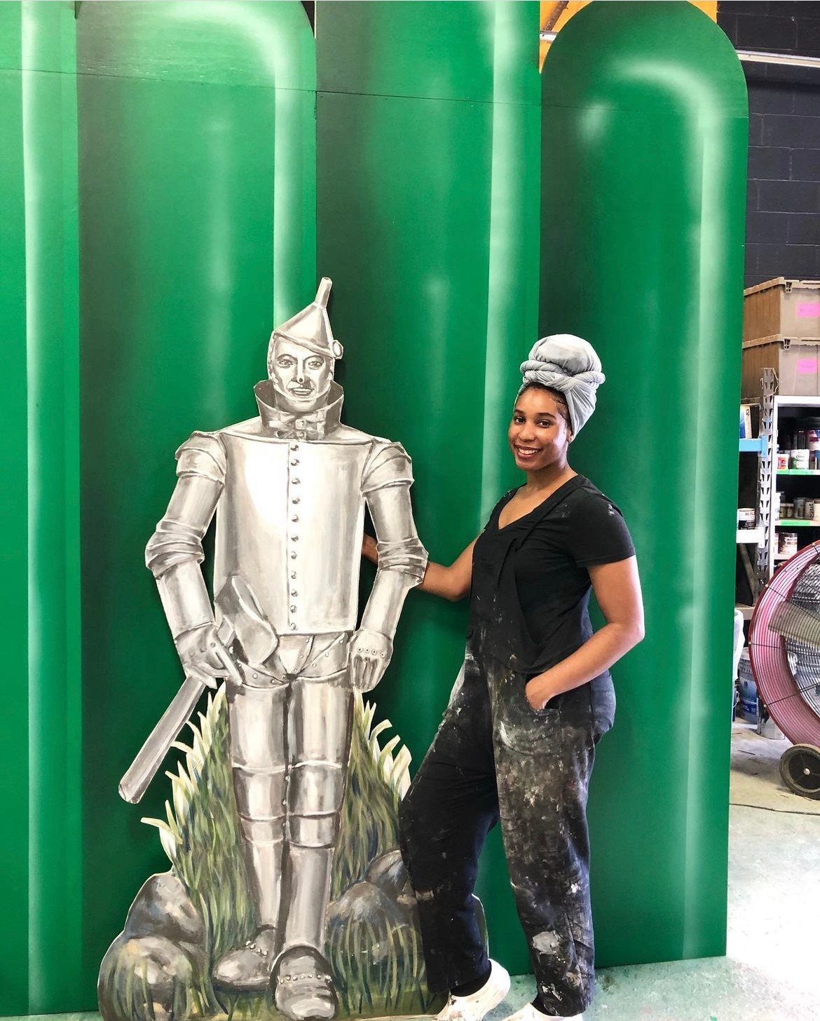 Woman with a hairnet standing next to a metallic Tin Man statue in front of large green structures, smiling, with a workshop background.