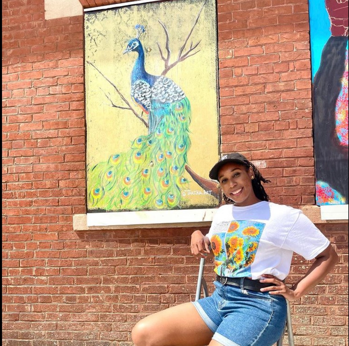 Young woman smiling, sitting outdoors in front of a brick wall with colorful artwork, pointing at a peacock painting.
