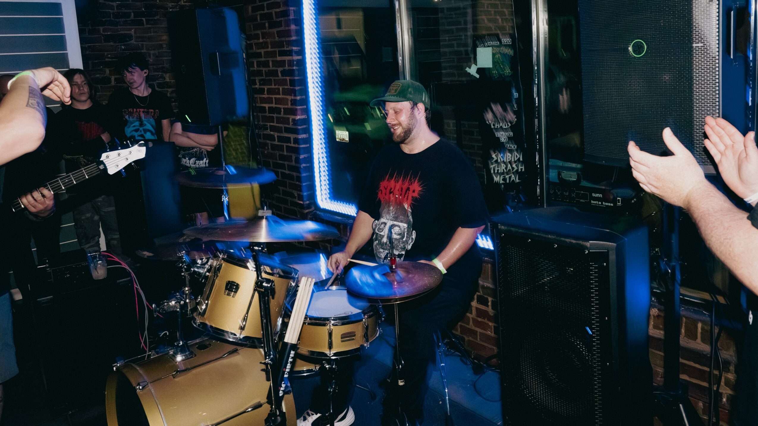A man plays drums in a band with other musicians, in a dimly lit venue with blue lighting.