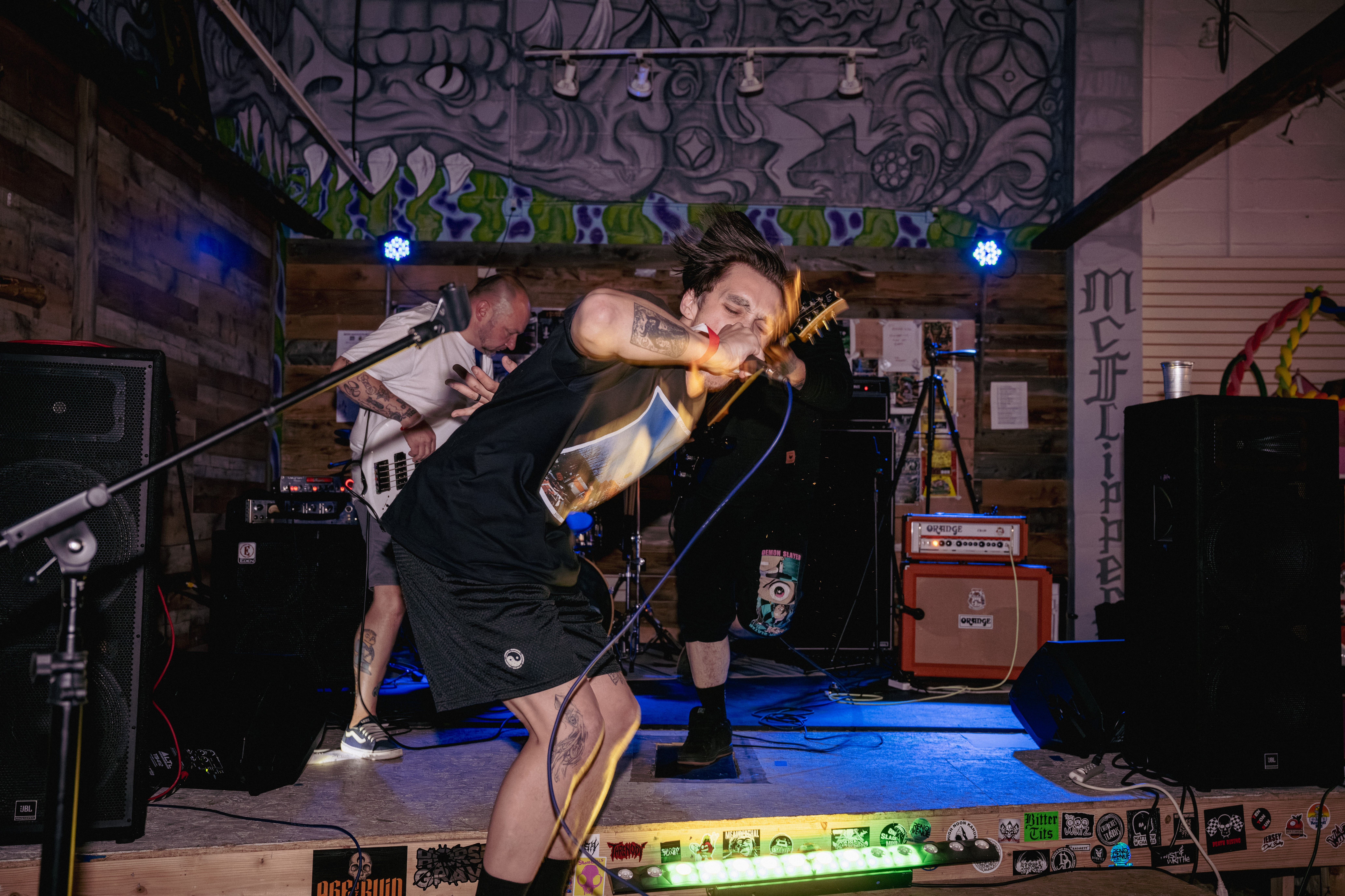 Person singing into a microphone on stage with band members and musical equipment, colorful background, performing live.