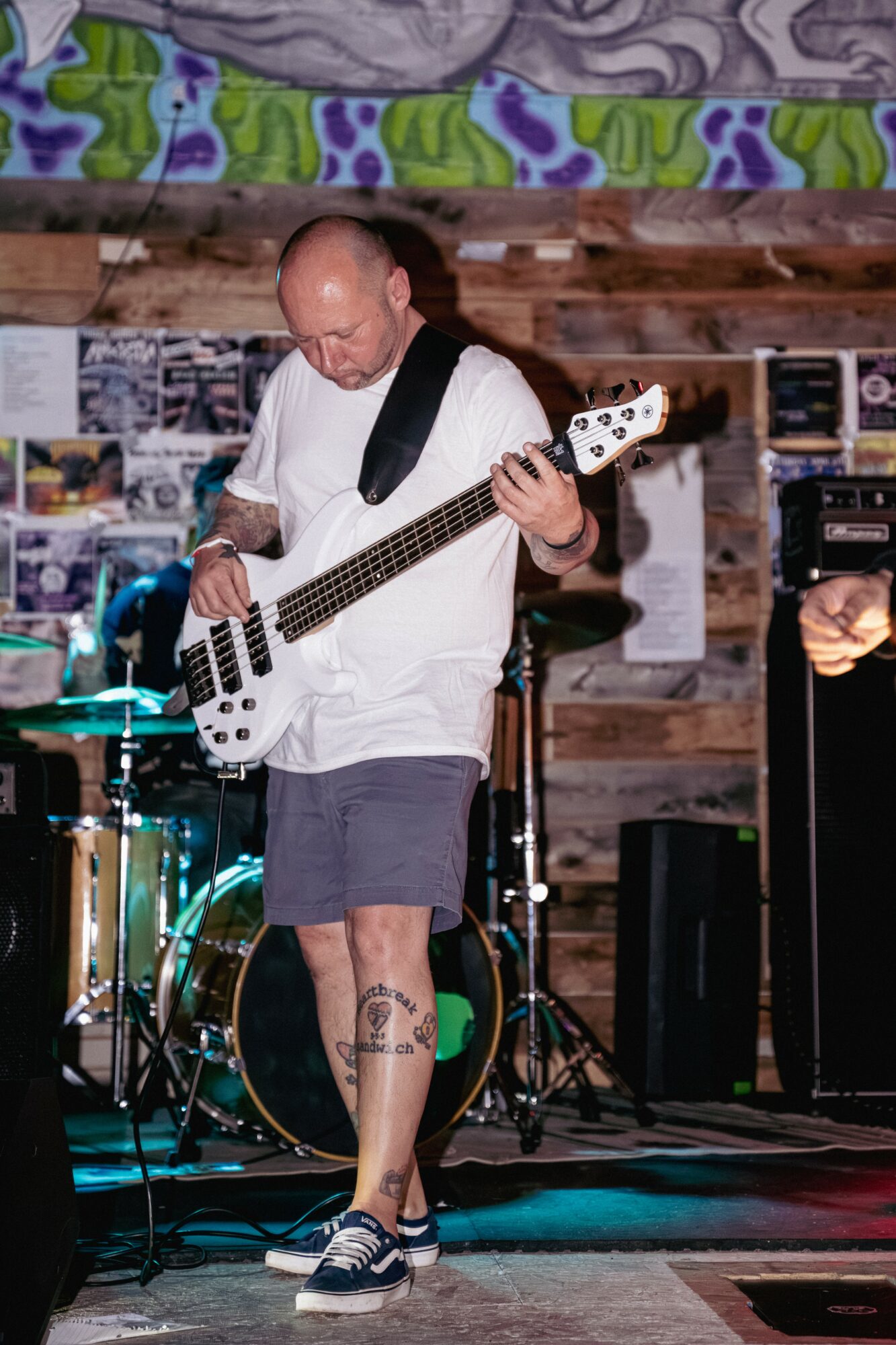 Bald man playing bass guitar on stage with wooden background and colorful mural above, wearing white shirt and shorts.