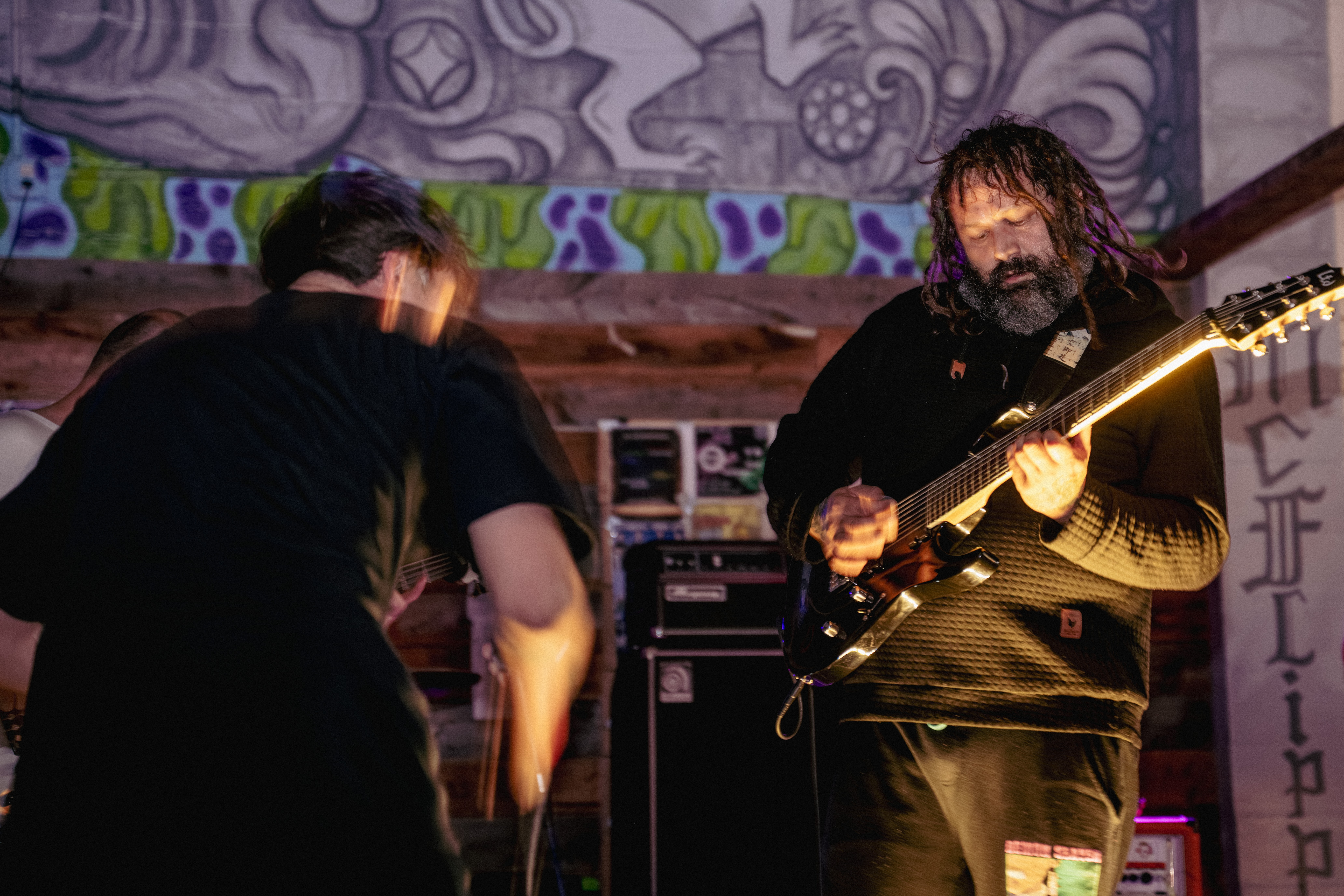 Musician playing electric guitar on stage with colorful mural background, person in black shirt in foreground, stage equipment visible.