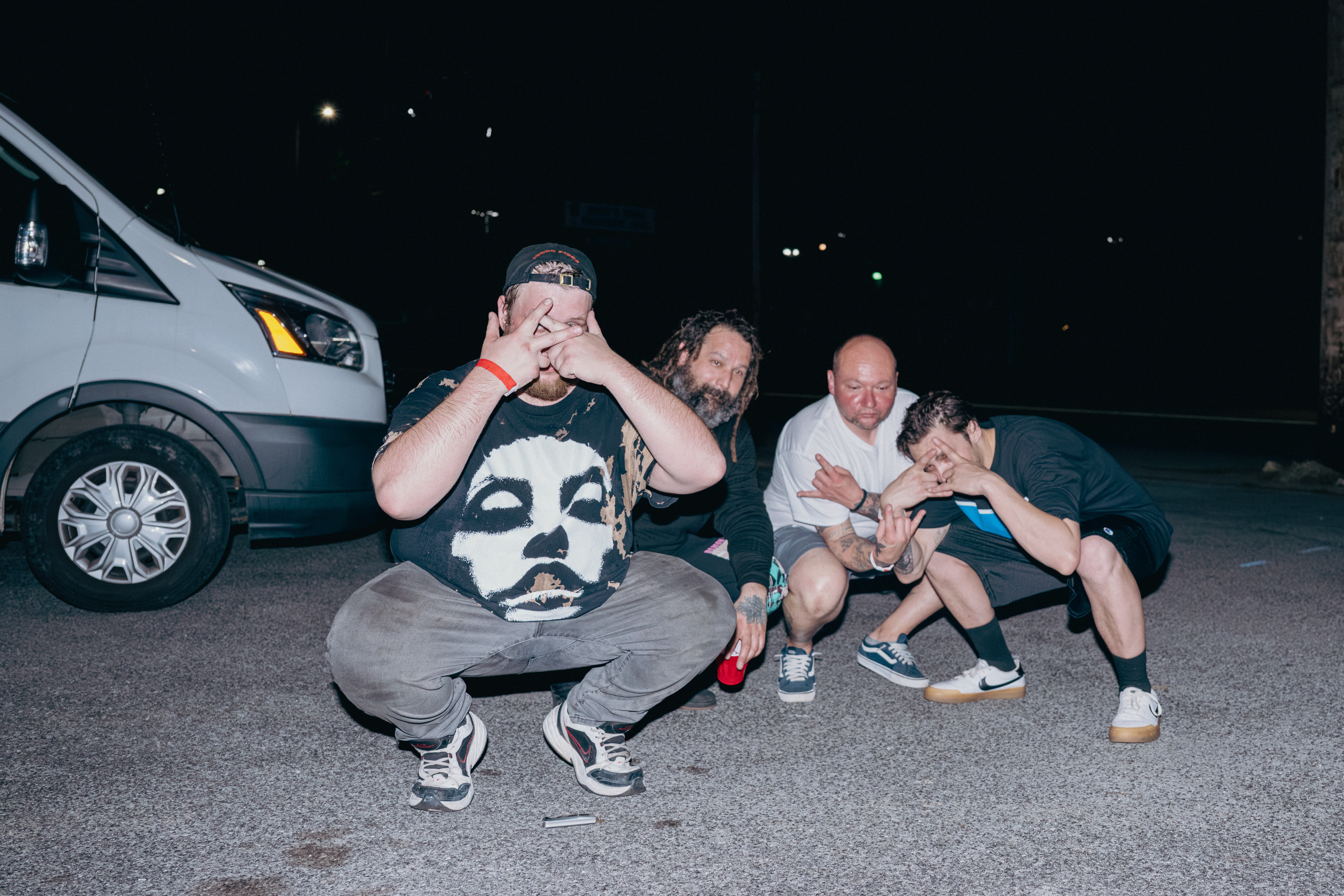 Four people crouching on the ground at night, with a vehicle and dark background behind them.