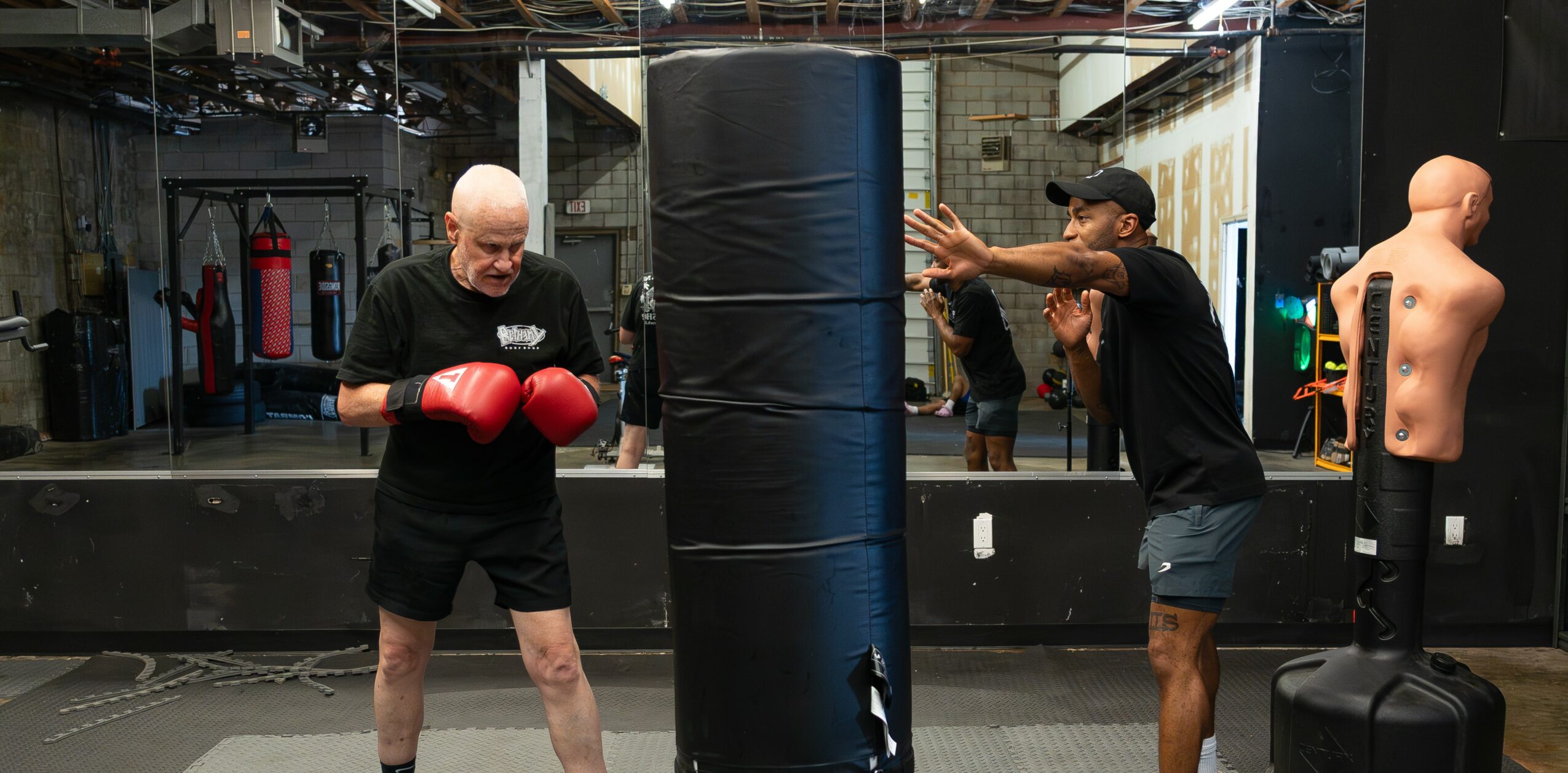 Two men training with a punching bag in a gym, one wearing red gloves and the other coaching.