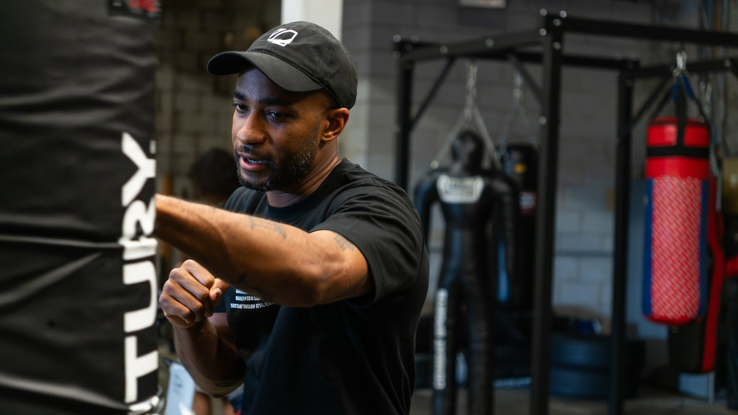Man in black shirt and cap training in gym with punching bag and equipment behind him.