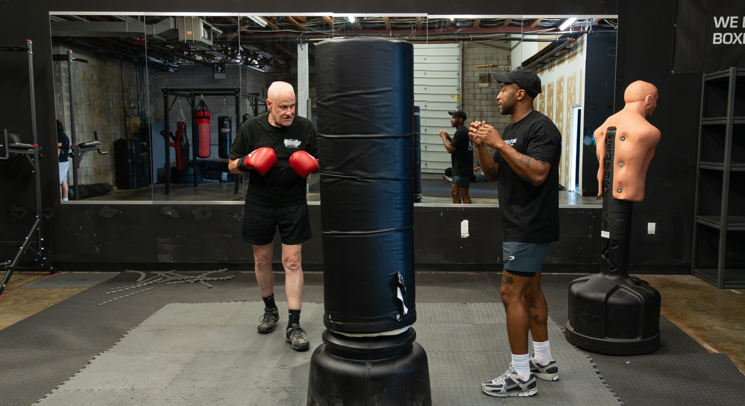 Two men in a boxing gym, one with boxing gloves and the other holding a camera, near a punching bag and training equipment.