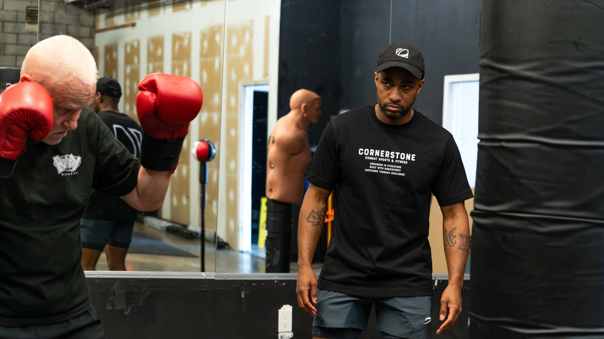 Boxing gym with people training, some wearing gloves, and a trainer observing. Equipment and mirrors are visible in the background.