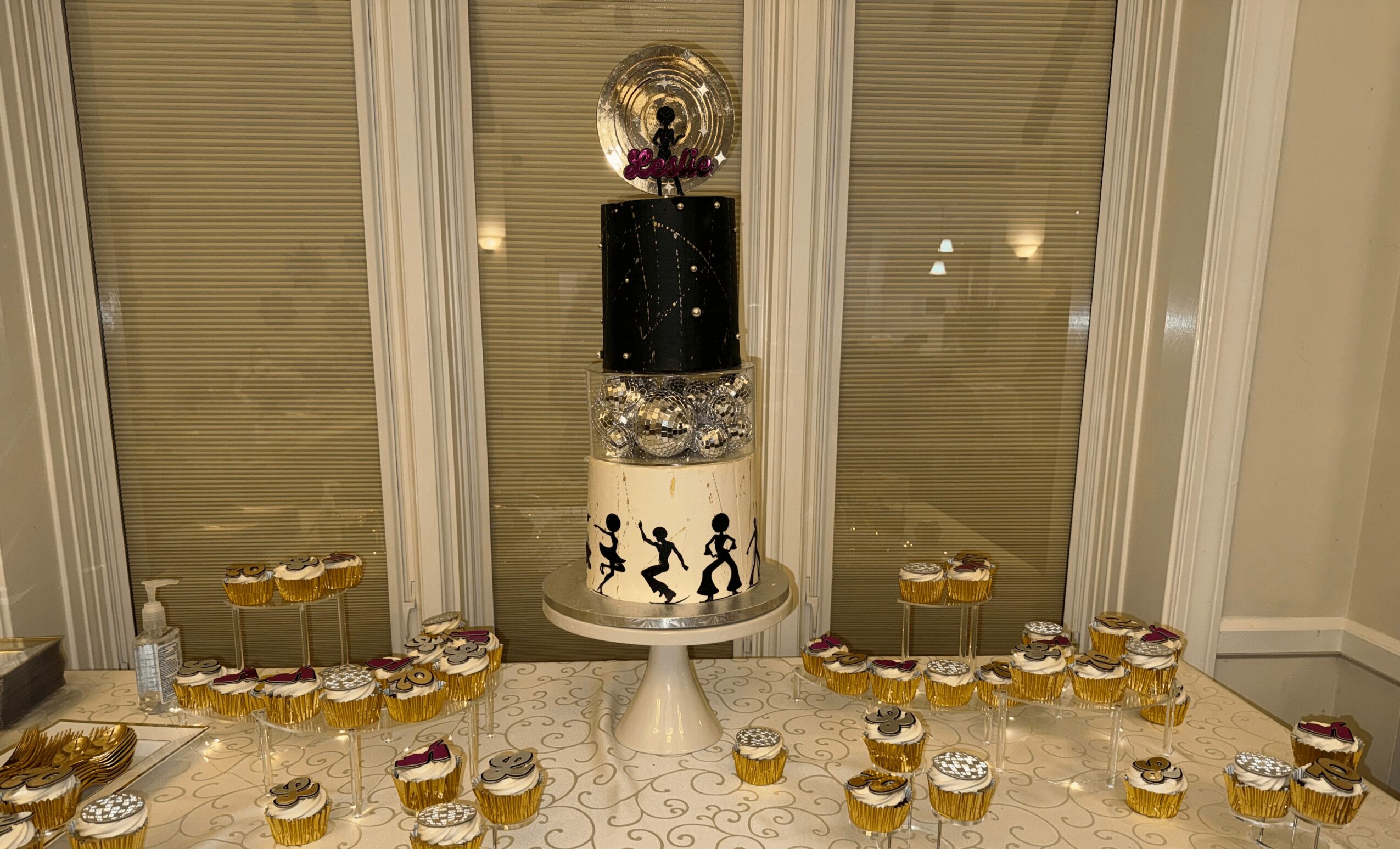 A tall cake with a black top, clear middle, and white base, decorated with dancing silhouettes, surrounded by cupcakes on a table.