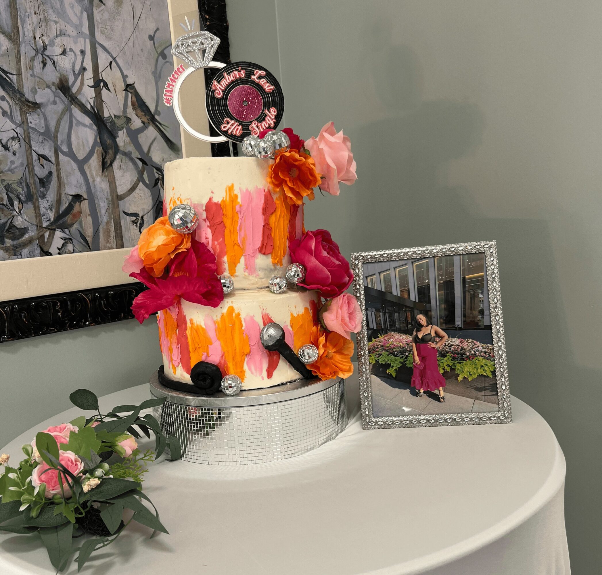 Two-tiered cake decorated with colorful brushstrokes, flowers, and metallic accents, placed on a white table with a framed photo nearby.