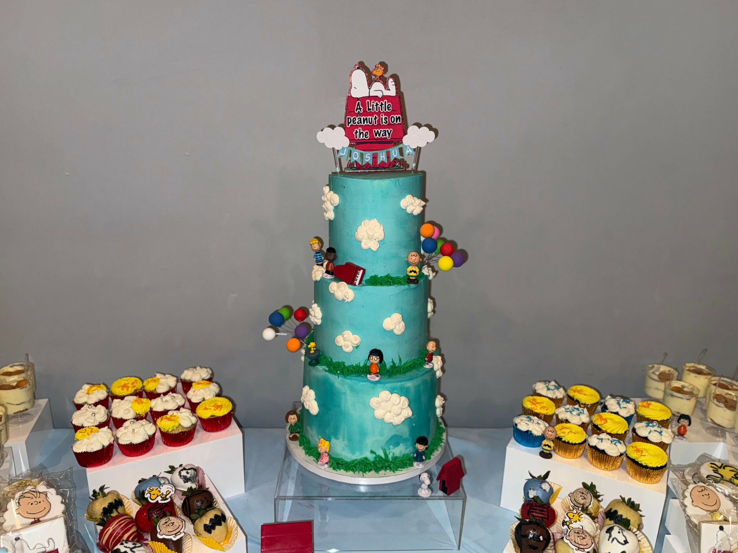 Three-tiered cake with clouds and characters, surrounded by cupcakes on a table, against a plain wall.