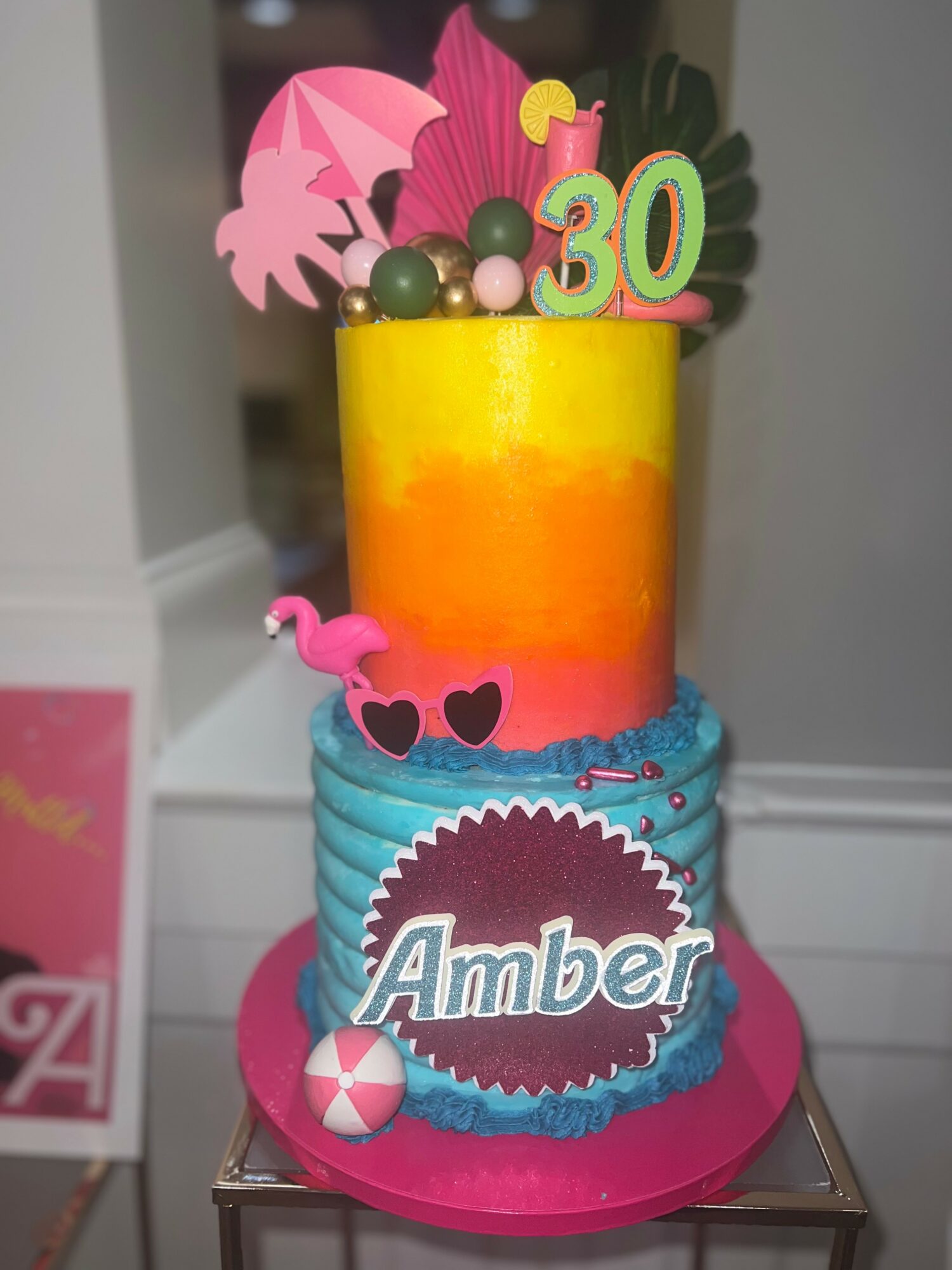 Two-tiered birthday cake with colorful decorations, pink flamingo, and a green number 30, on a pink base.