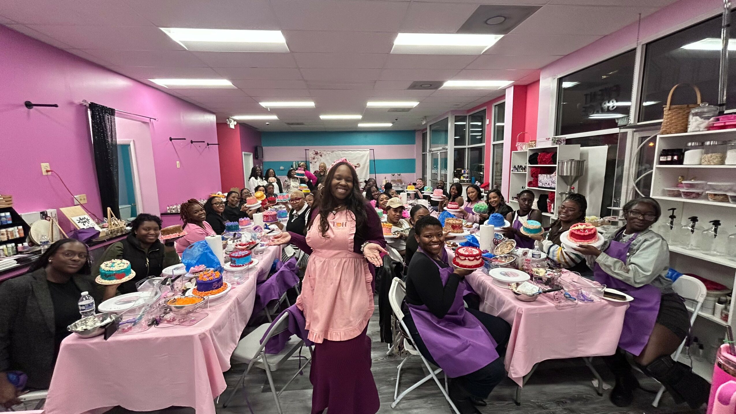 A woman in a pink dress stands in a decorated room with children seated at tables, celebrating with cakes and gifts.