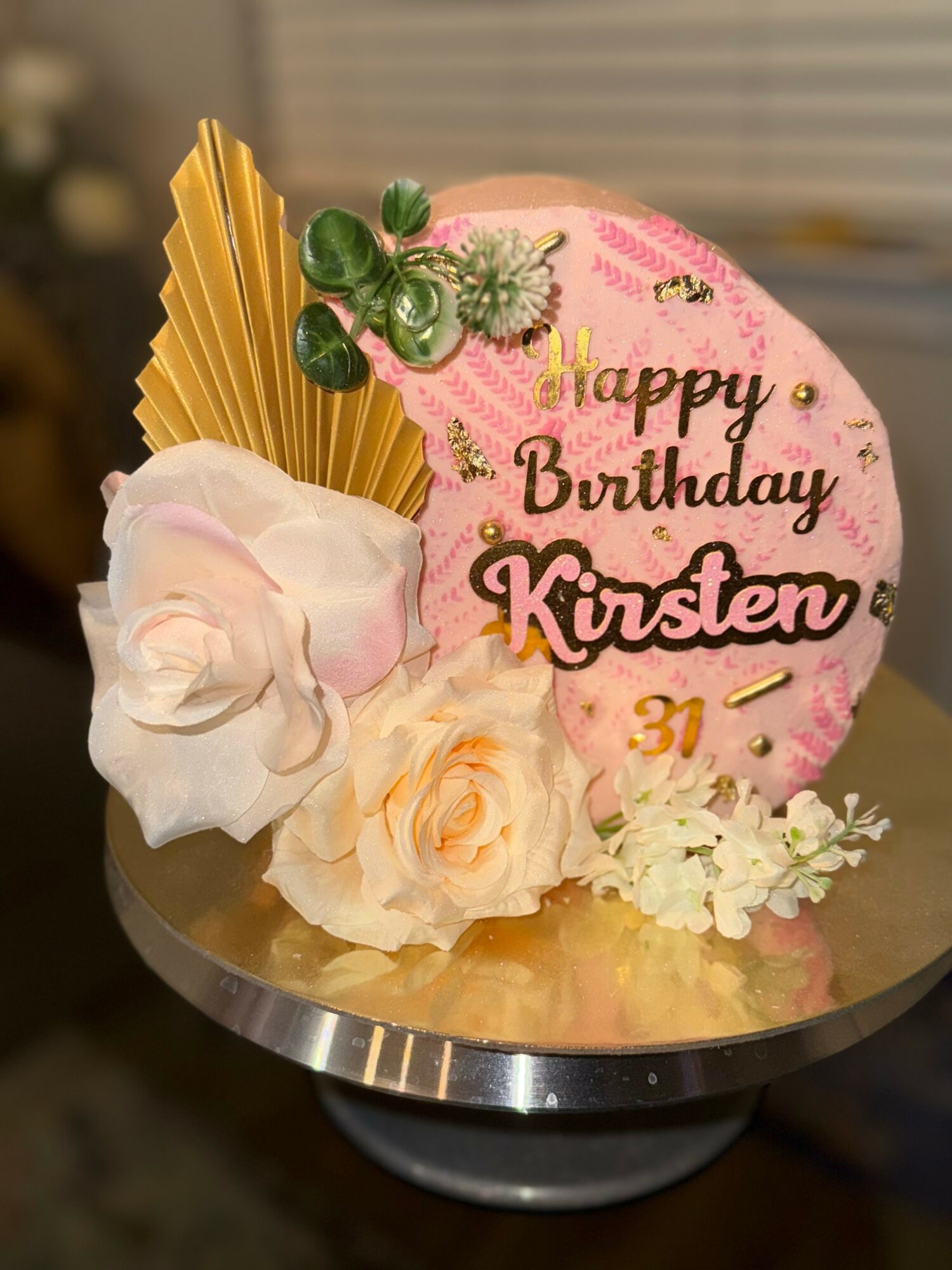 Round birthday cake with pink and white frosting, decorated with flowers, gold accents, and a message for Kirsten's 31st birthday.