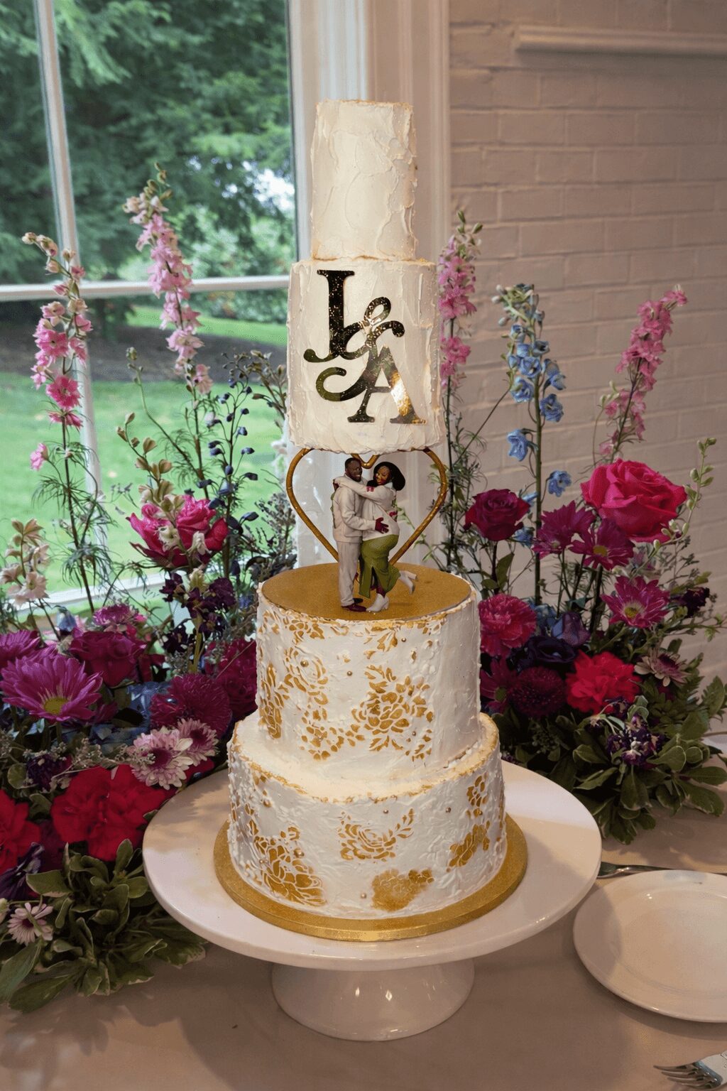 Three-tiered wedding cake with gold accents, topped with a cake topper and surrounded by colorful flowers, near a window.