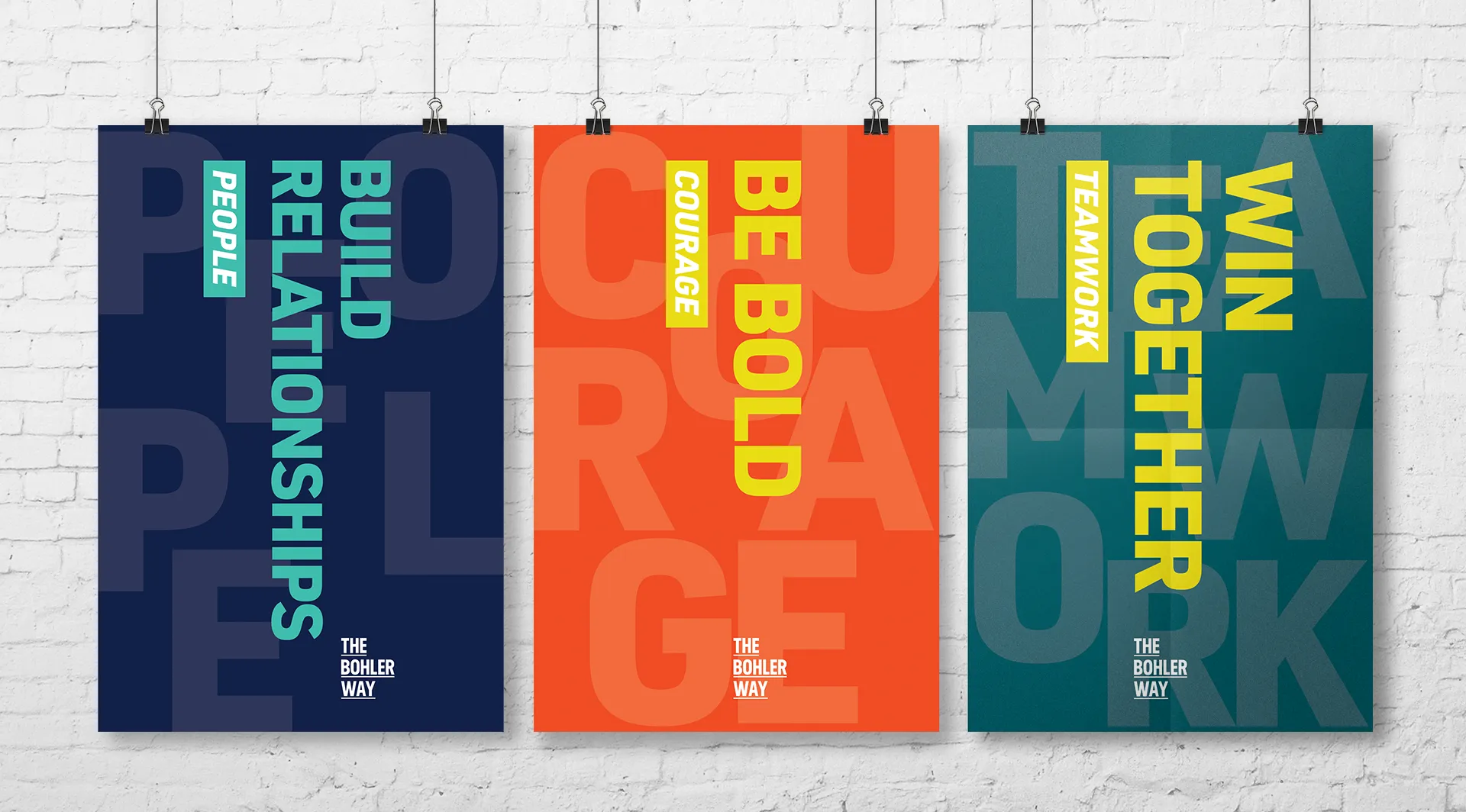 Three hanging posters with motivational messages about building relationships, being bold, and winning together, on a white brick wall.