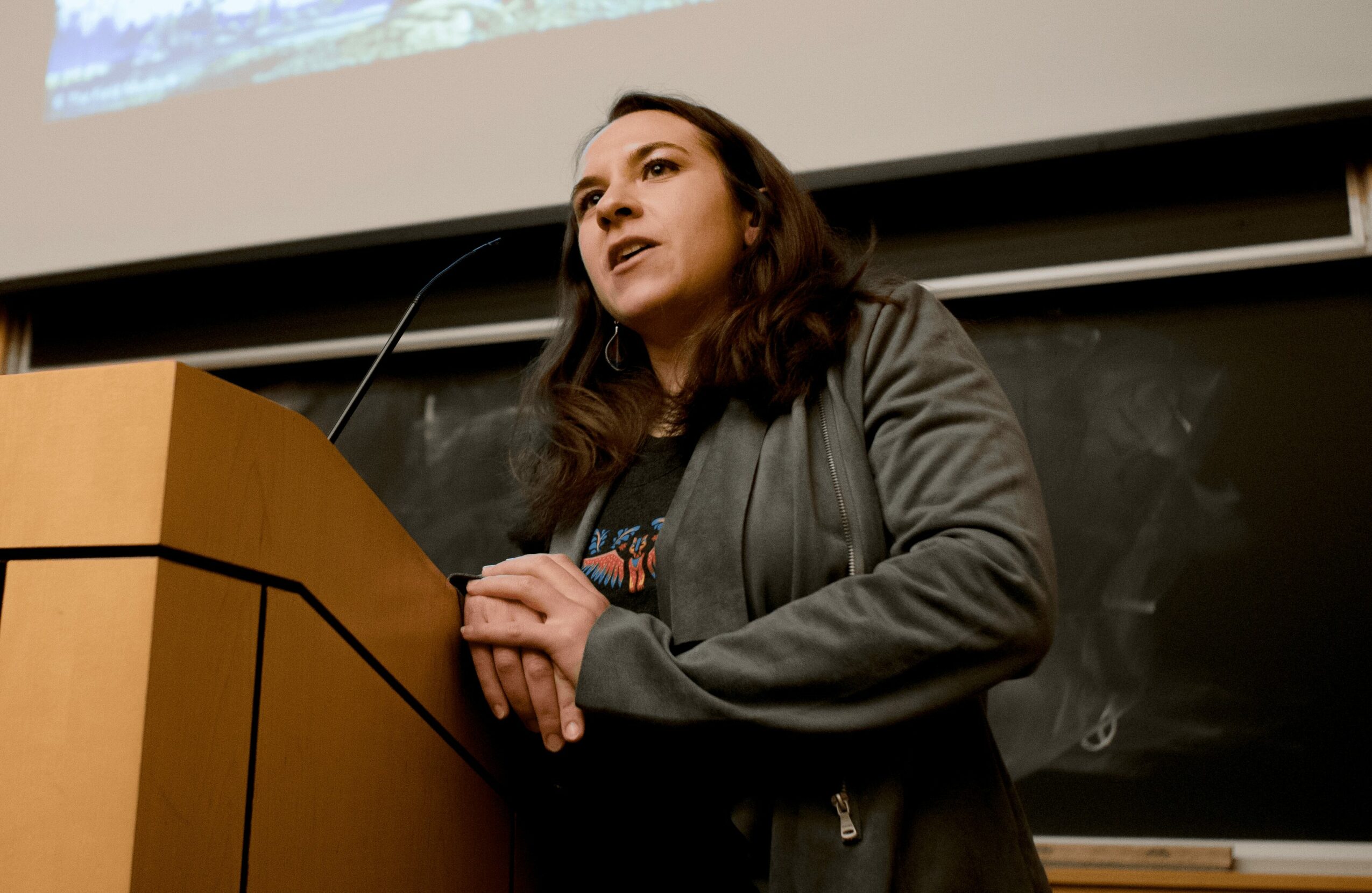Woman speaking at a podium in a classroom with a blackboard behind her.