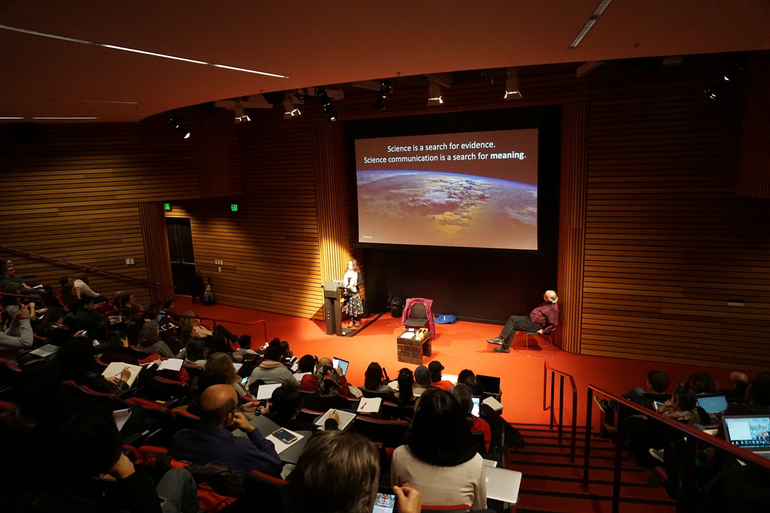 Audience seated in a conference room watching a presentation on a large screen, with a speaker at a podium and a person sitting nearby.