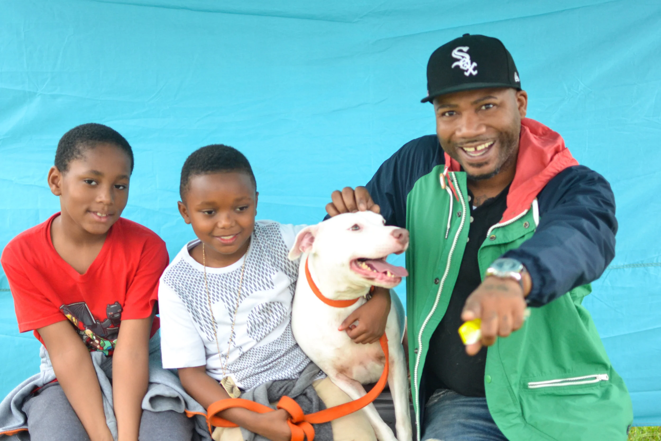 Man with two children and a white dog outdoors against a blue background, smiling and holding a yellow object.